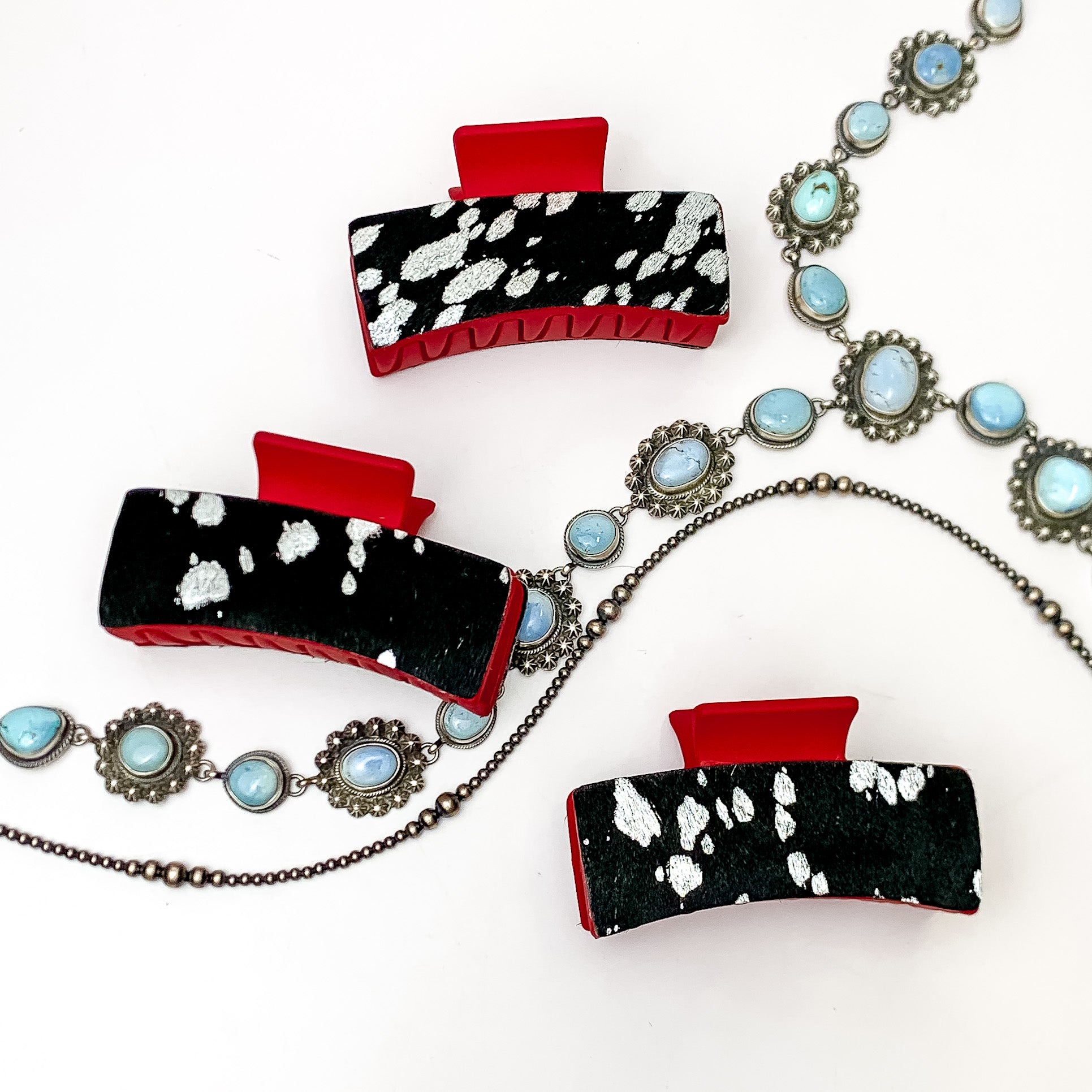 Pictured are three red hair clips with black and silver hide patches. These earrings are pictured on a white background with silver necklaces under the clips. 