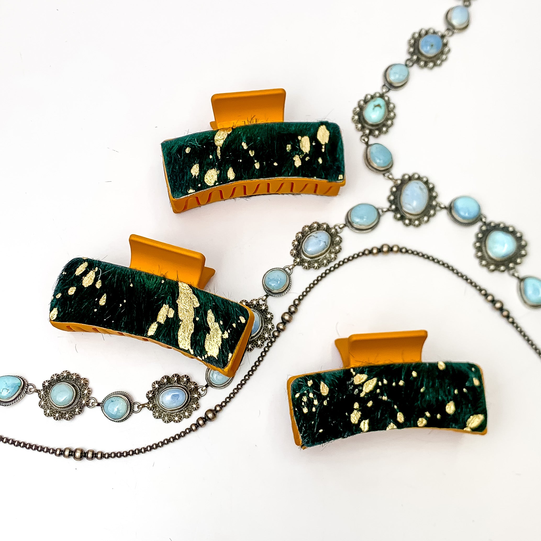 Pictured are three orange hair clips with green and gold hide patches. These earrings are pictured on a white background with silver necklaces under the clips. 