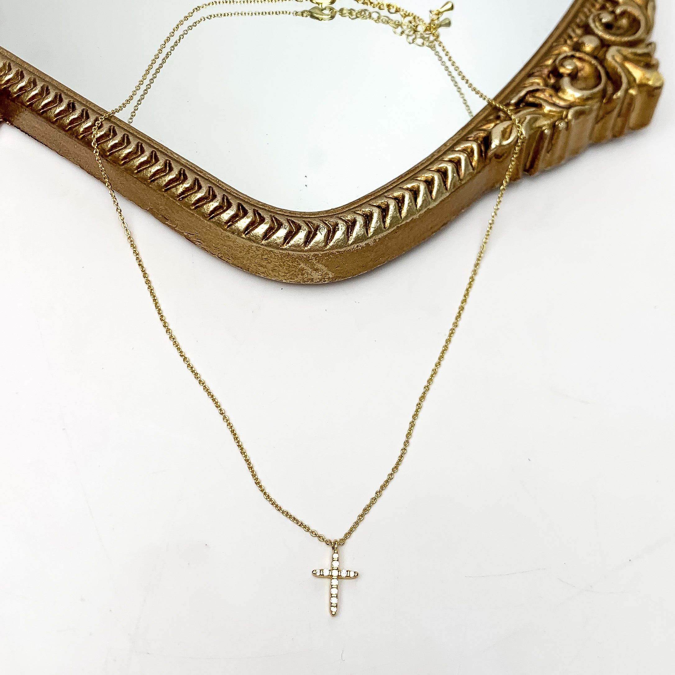 Gold Tone Glory Chain Necklace With Clear Crystal Cross. This necklace is pictured on a white background with part of it on a gold trimmed mirror.