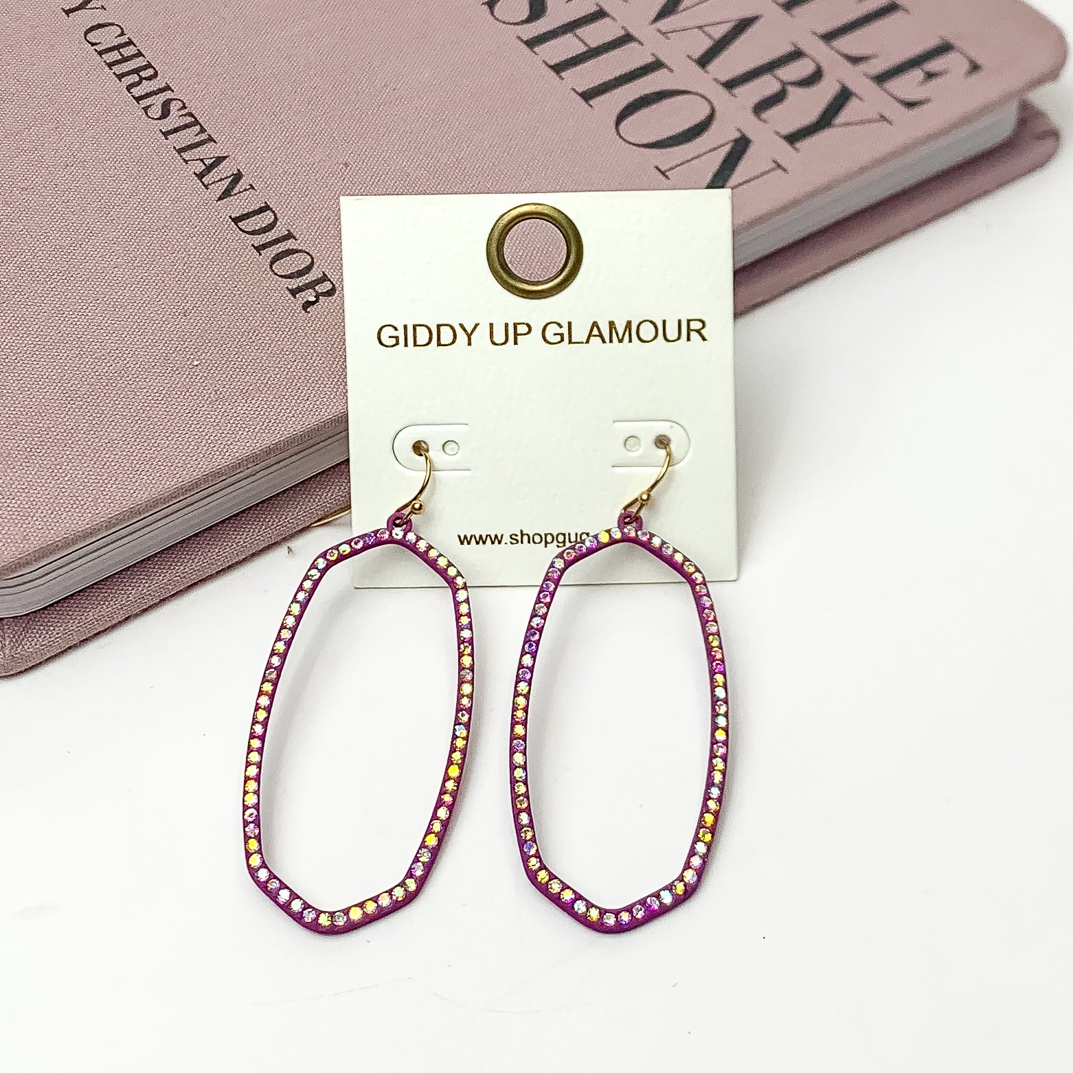 Sparkle Girl Open Oval Earrings in Purple. These earrings are pictured laying against a pink book with a white background.