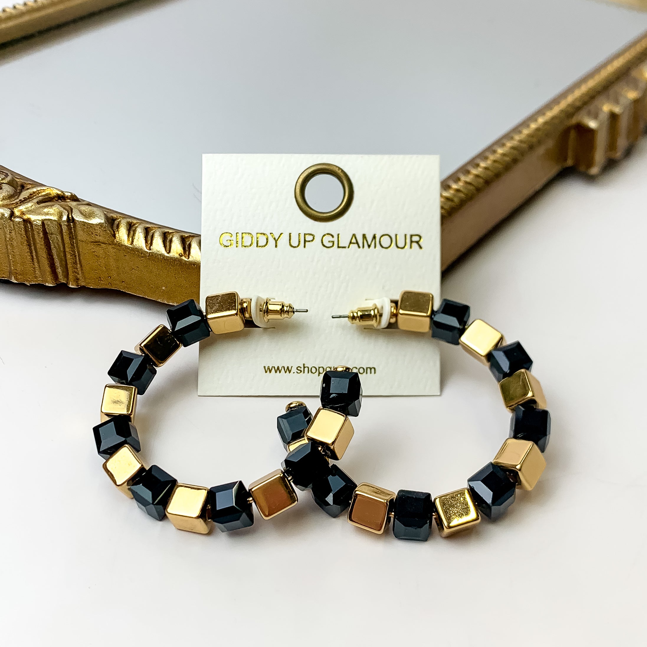 A pair of black and gold cubed hoop earrings on an ivory earring card. These earrings are placed against a gold mirror with a white background.