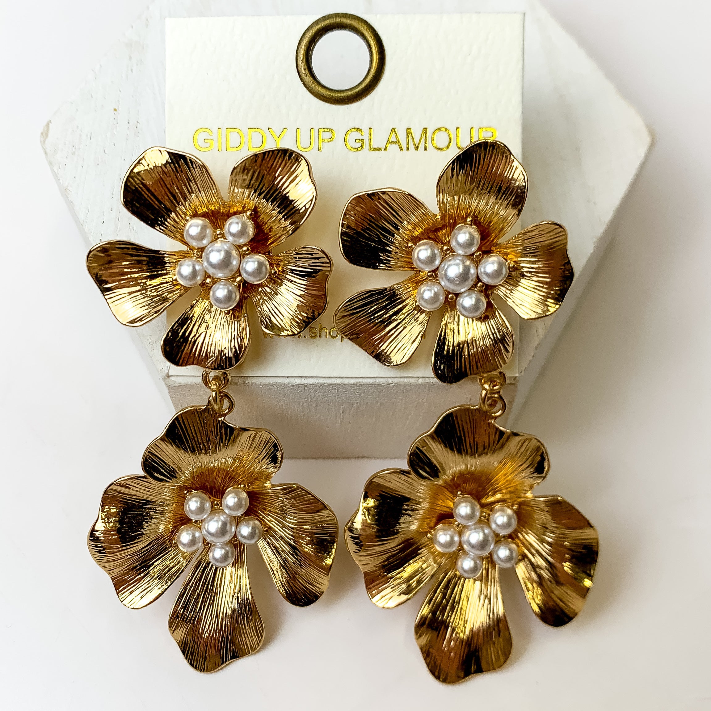 These Gold Tone Two Tiered Flower Earrings with a pearl center are placed on an ivory earring card. They are pictured on a white hexagon for lengthening purposes with a white backbround.