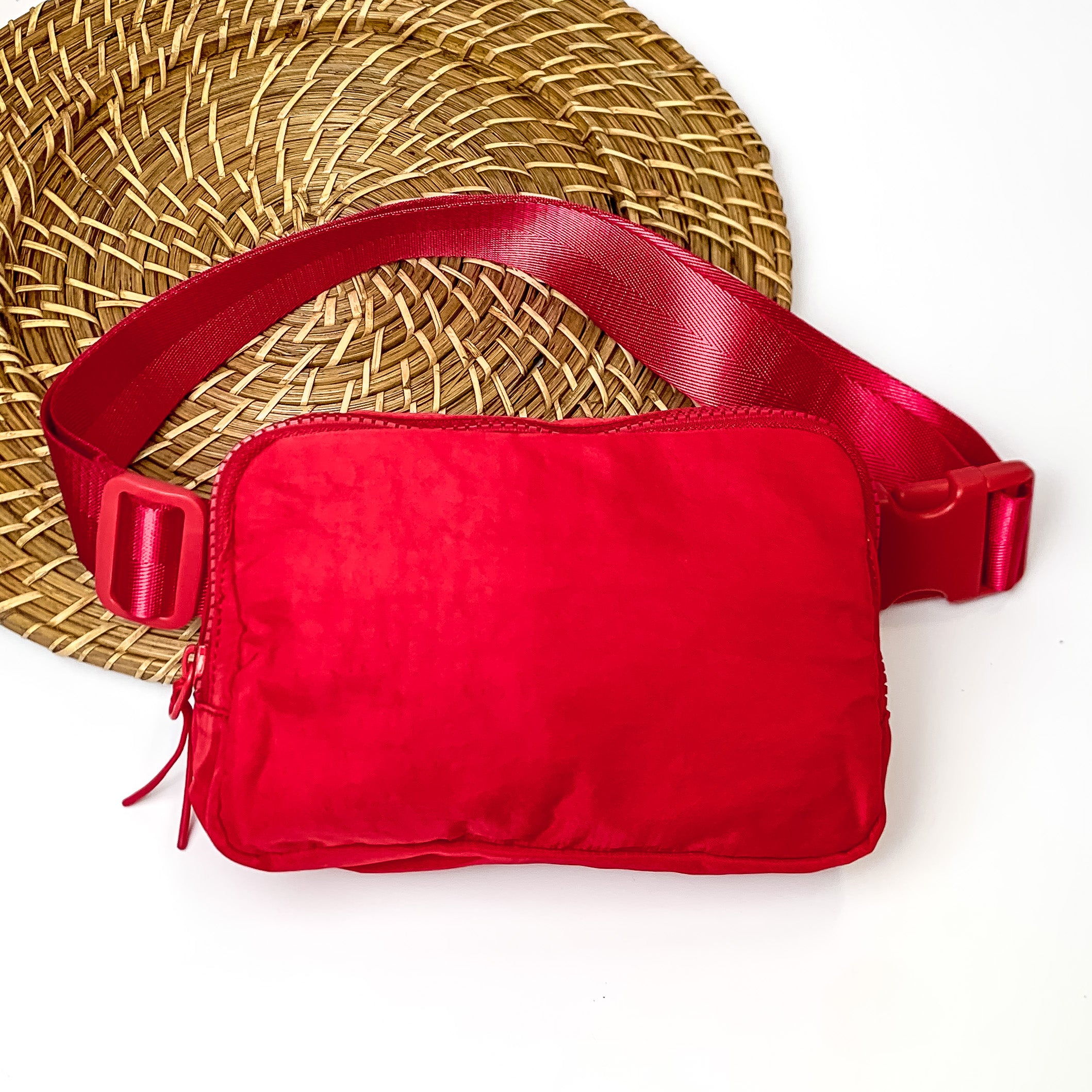 Pictured is a rectangle fanny pack with a top zipper with tassel in red. This bag also includes a red strap and red accents. This bag is pictured on a white and brown patterned background.