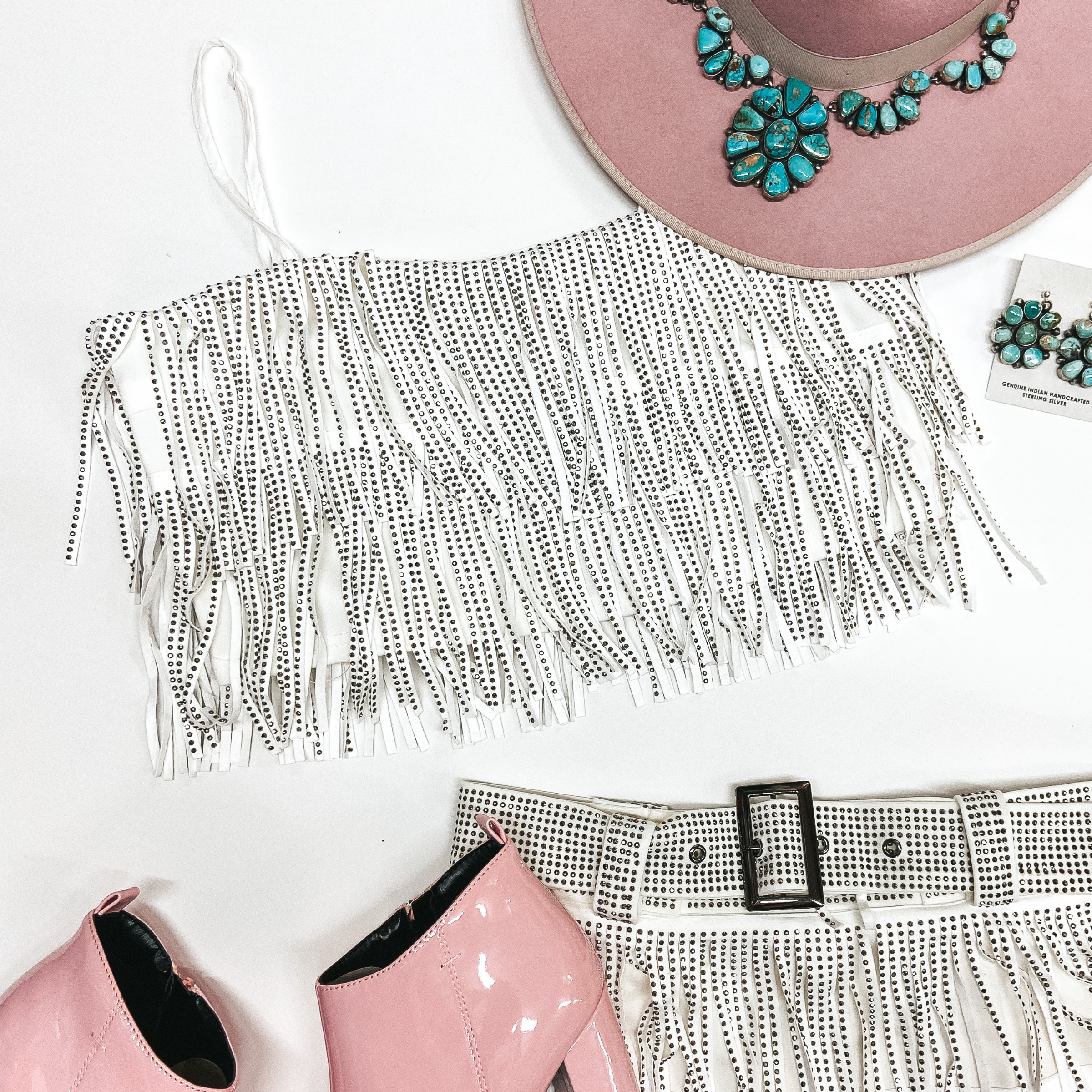 A white crystal fringe crop top with adjustable spaghetti straps pictured on a white background with a pink hat, pink booties, and turquoise jewelry.
