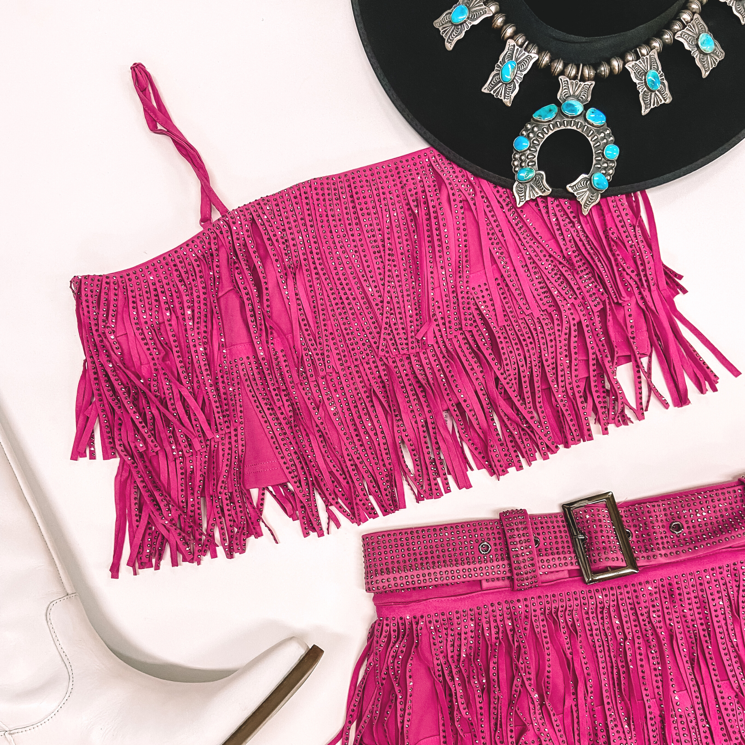 A pink crystal fringe crop top with adjustable spaghetti straps pictured on a white background with white boots, a black hat, and turquoise jewelry.
