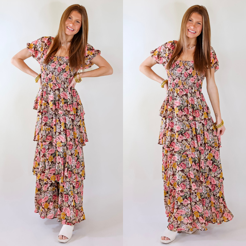 Model is wearing a tiered maxi dress with balloon sleeves. The dress has a floral print with brown, pink, and yellow. Model has paired the dress with white open toe heels, pearl and gold tone bracelets, and a gold tone necklace.