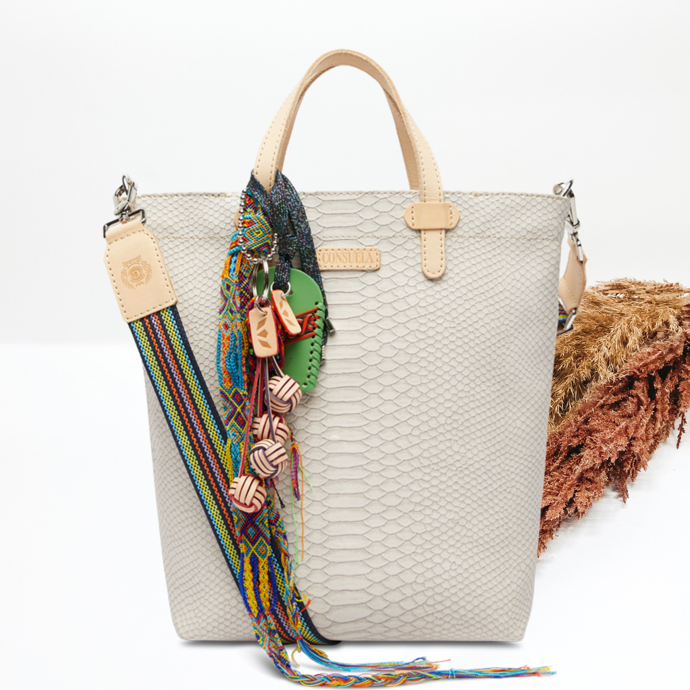 Pictured on a white background is an essential tote bag that has a white snake print. This purse includes a olive and tan charm, a striped purse strap, and light tan handles.