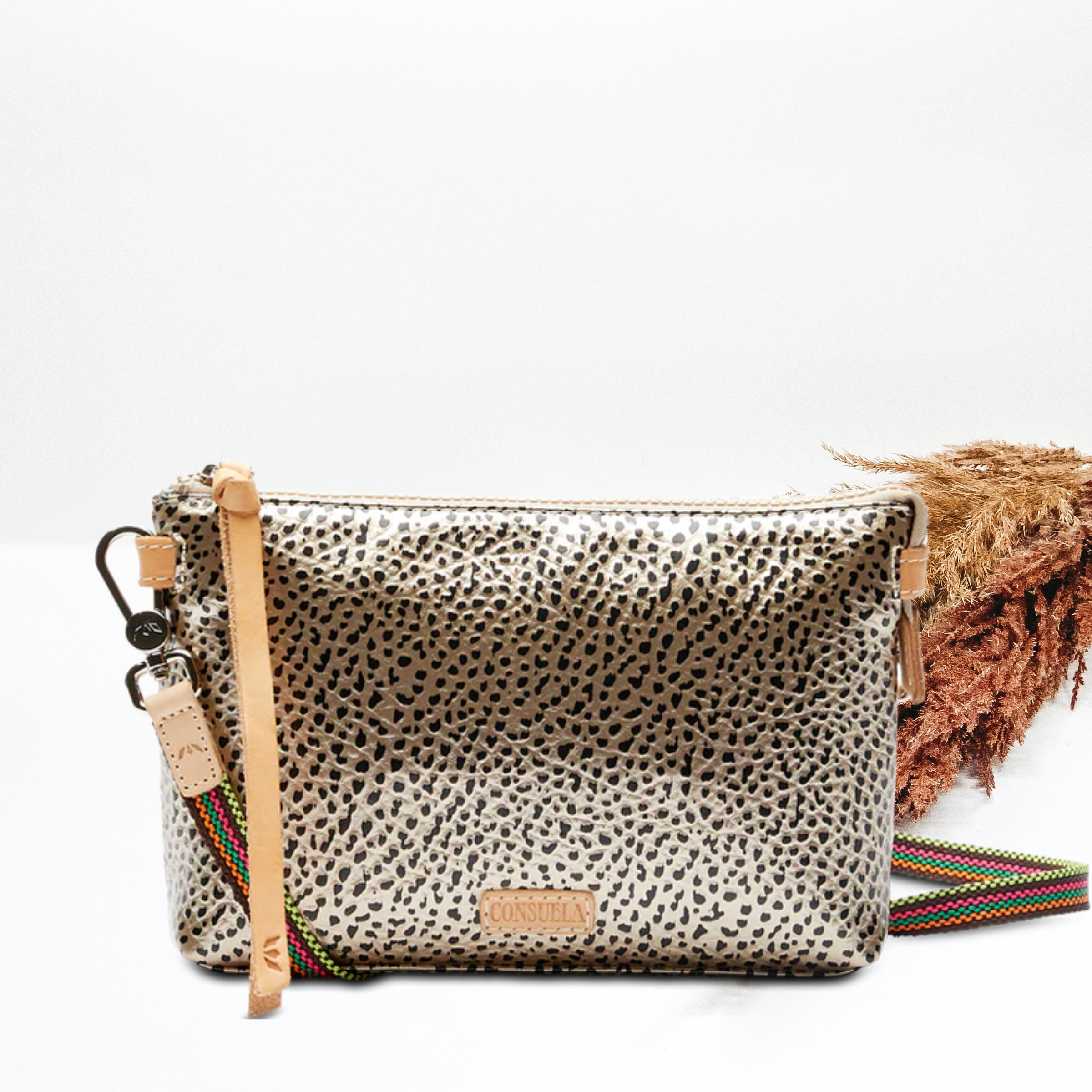 A gold metallic leather crossbody purse with leather detailing. Pictured on white background with a brown and tan pompous grass on the right hand side. 