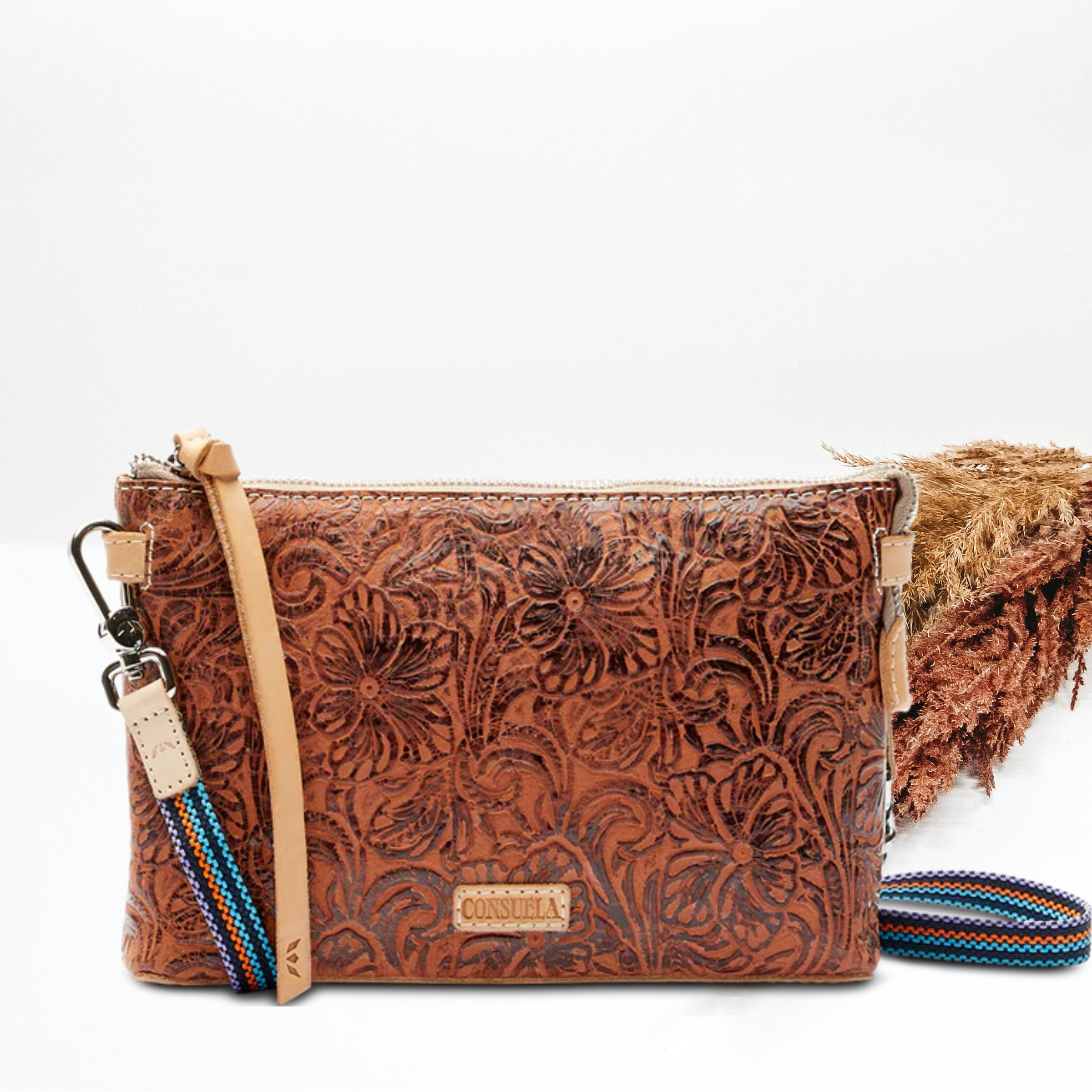 A light brown leather tooled purse with a crossbody strap. Pictured on white background with brown pompous. 