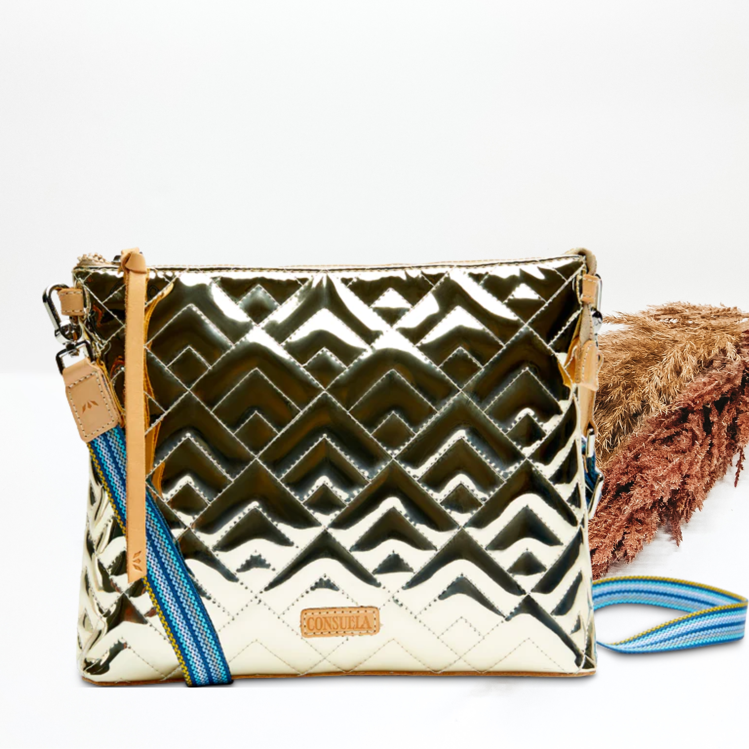 A gold metallic purse with a crossbody strap. Pictured on white background with brown pompous grass on the right side. 