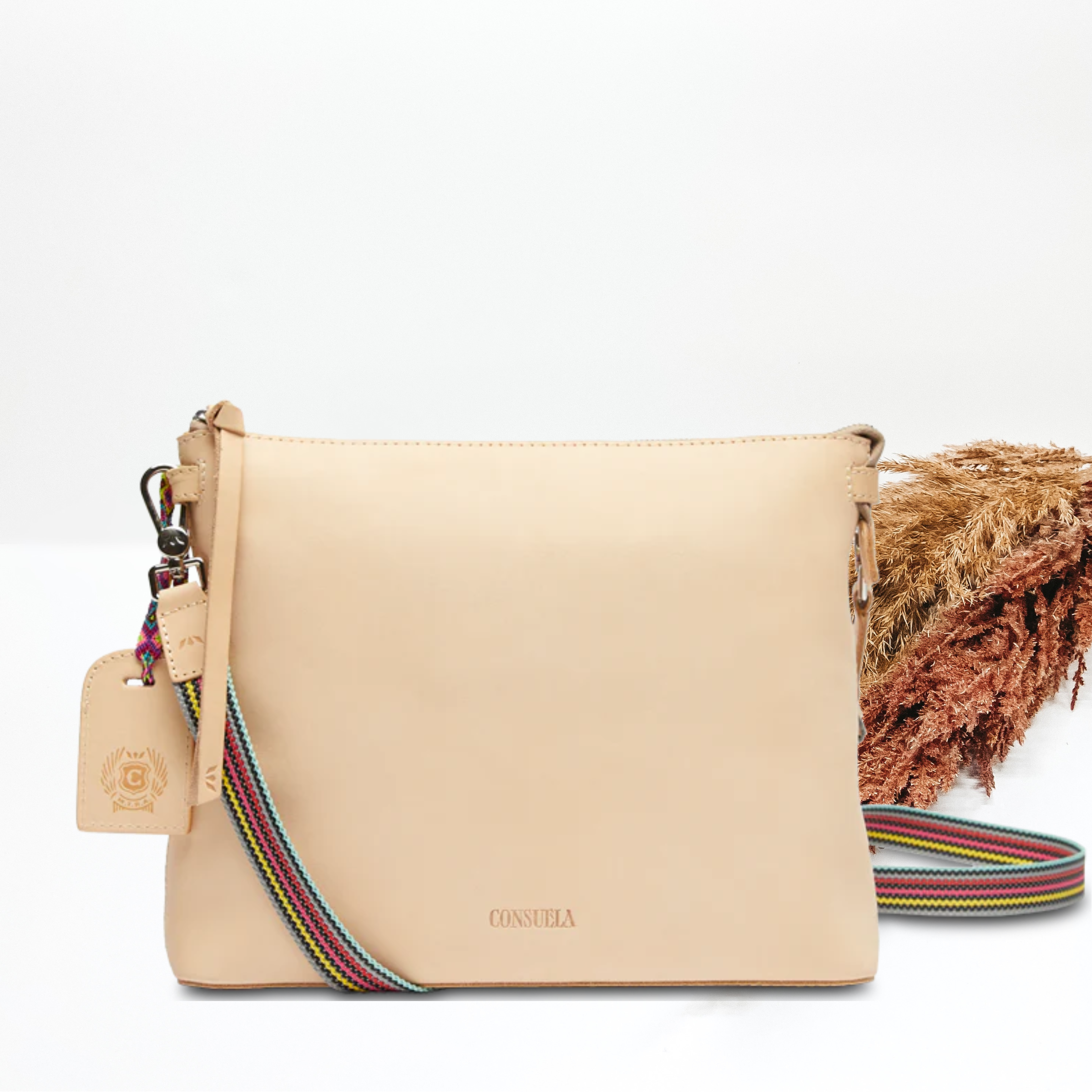 A light leather crossbody bag with a colorful strap. Pictured on white background with brown pompous grass on the right side.