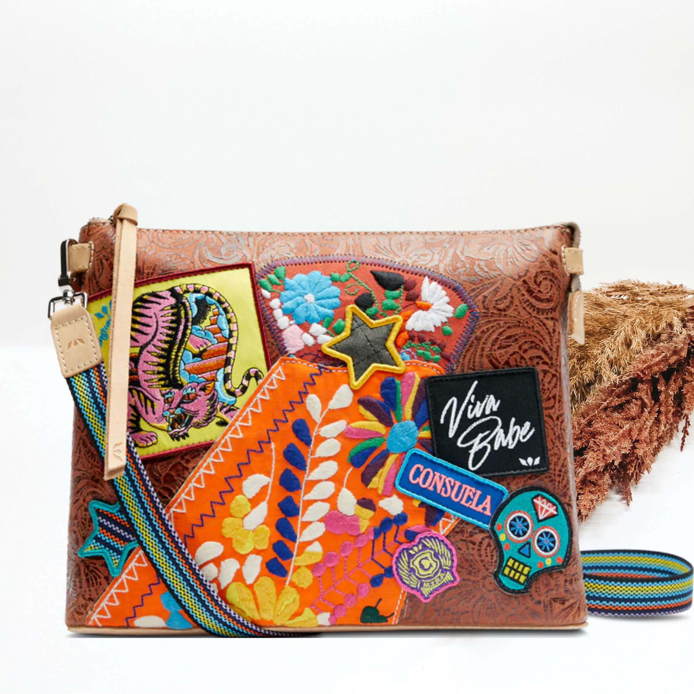 A leather tooled print purse with colorful patches and a crossbody strap. Pictured on white background with brown pompous grass on the right side. 