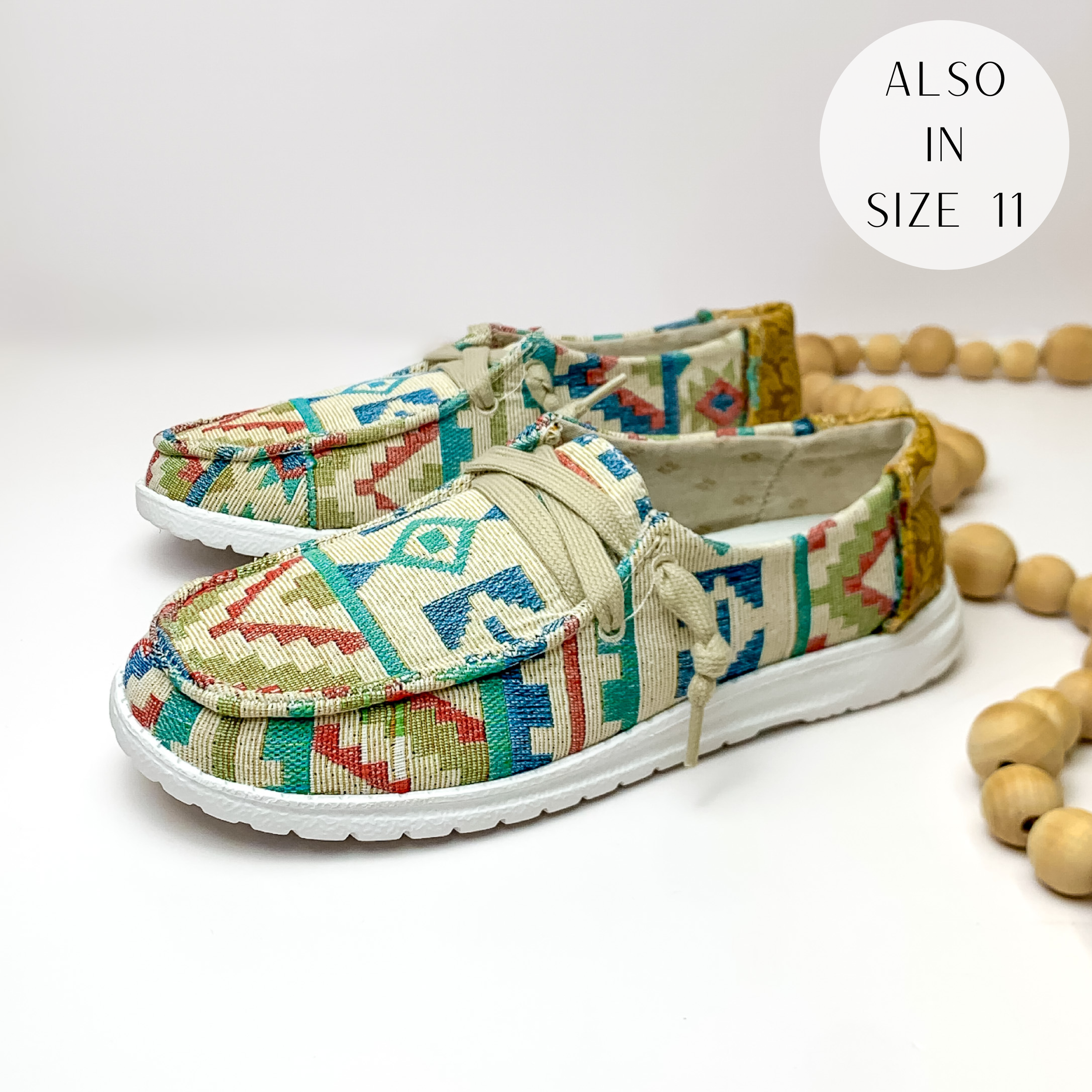 Pictured are a pair of multicolored tribal print canvas shoes with a white sole and beige laces. These shoes also include tan, leather tooled heels. These shoes are pictured on a white background with tan beads on the right side.