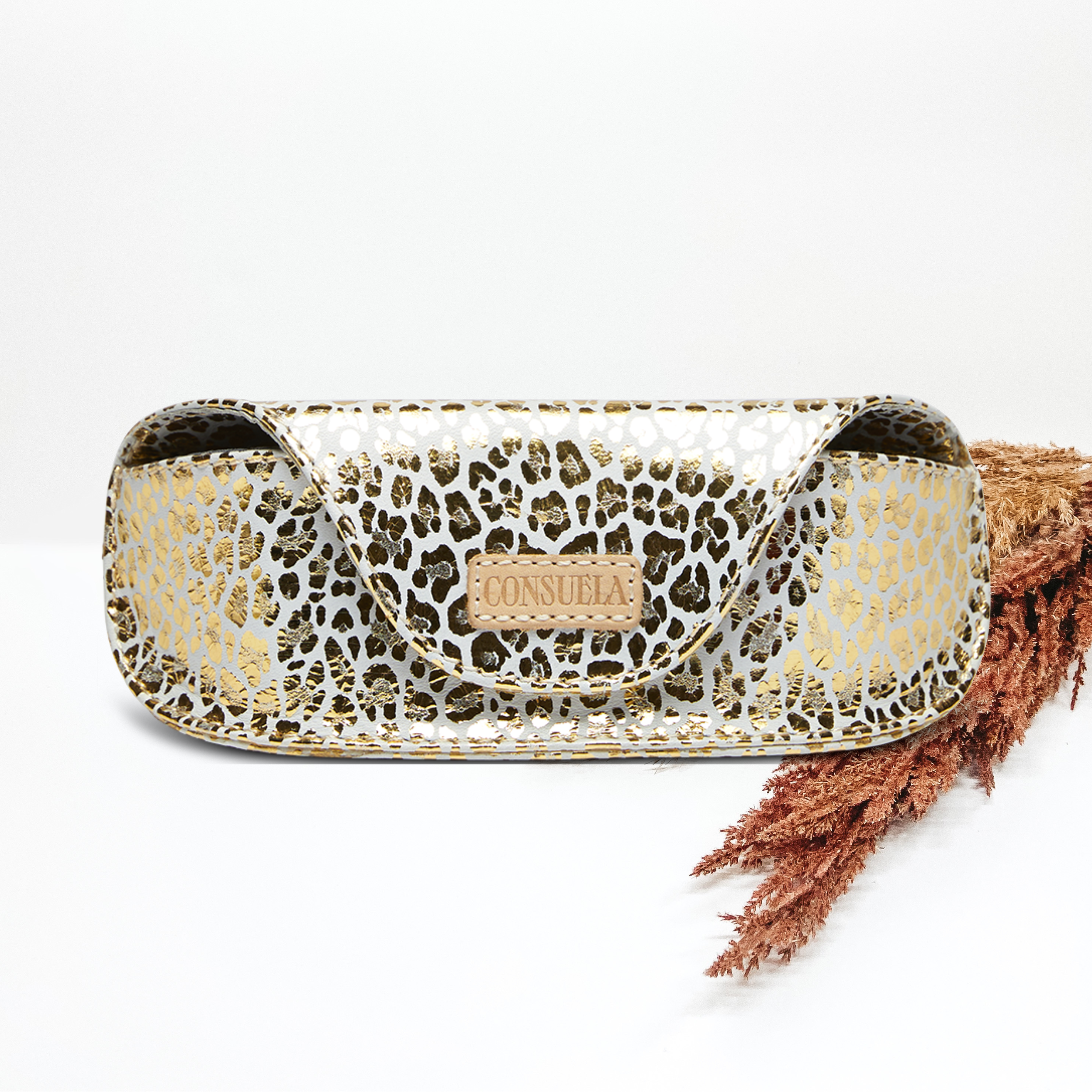 Pictured on a white background is a sunglasses case with a light tan leather patch that says "CONSUELA". This case is white with a metallic gold leopard print design. 