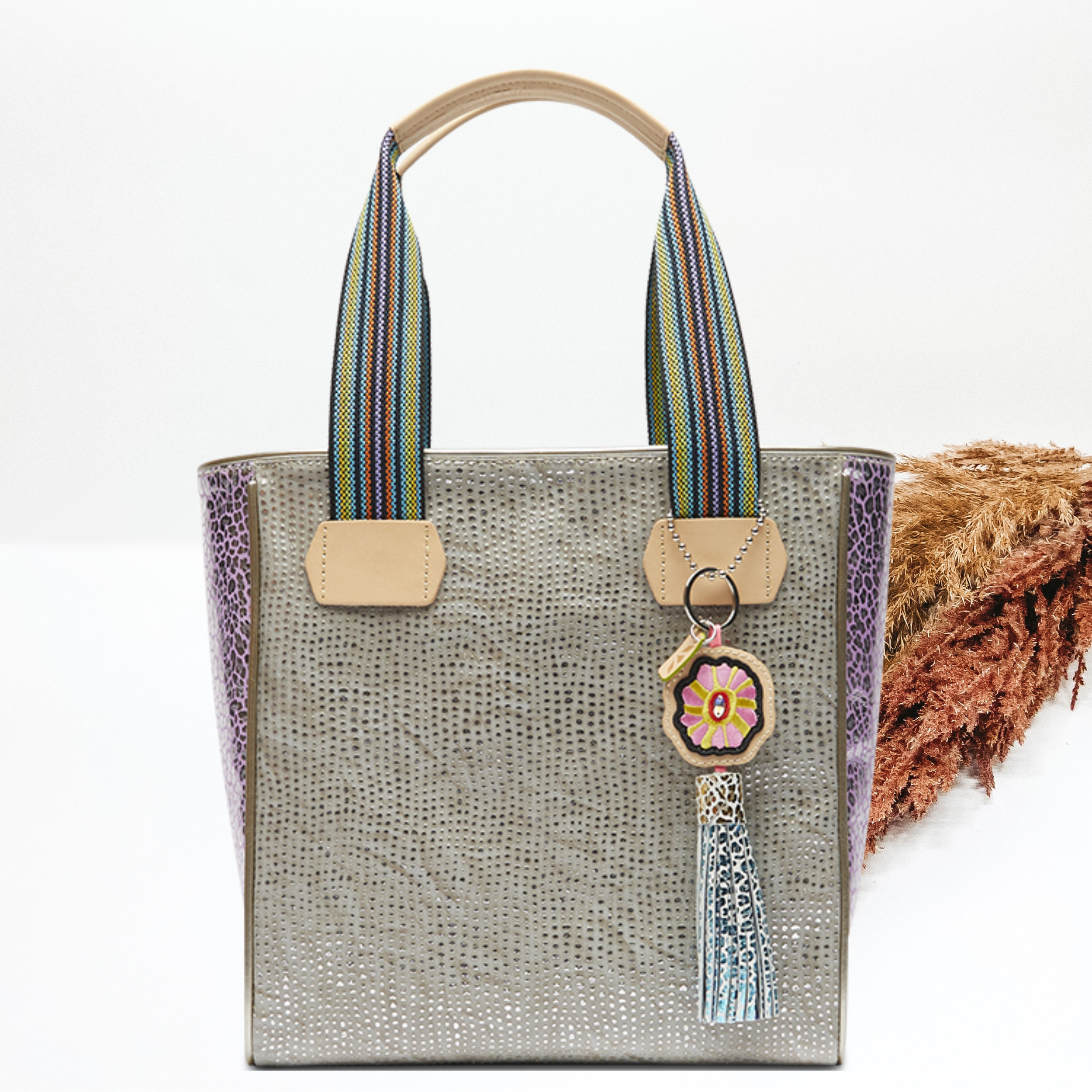 Pictured on a white background is a grey classic tote bag with woven straps and light tan handles. This bag includes a silver dotted print, purple leopard side panels, and a multicolored tassel charm. 