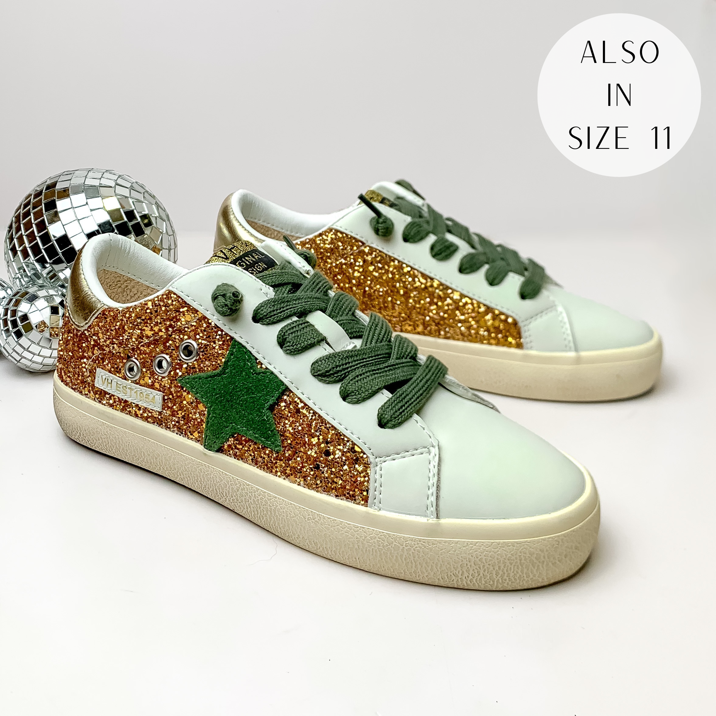 Pictured is a pair of ivory tennis shoes with gold and olive green detailing. These shoes include a gold shimmer heel, an olive green star on the side, and olive green laces. These shoes are pictured on a white background with disco balls on the left side