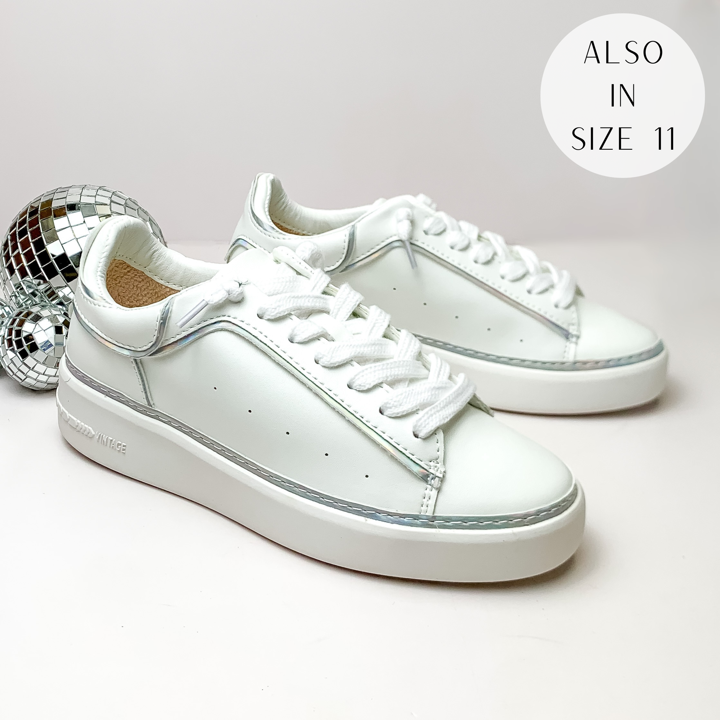Pictured is a pair of white tennis shoes with white laces and a silver iridescent outline. These shoes are pictured on a white background with disco balls on the left side