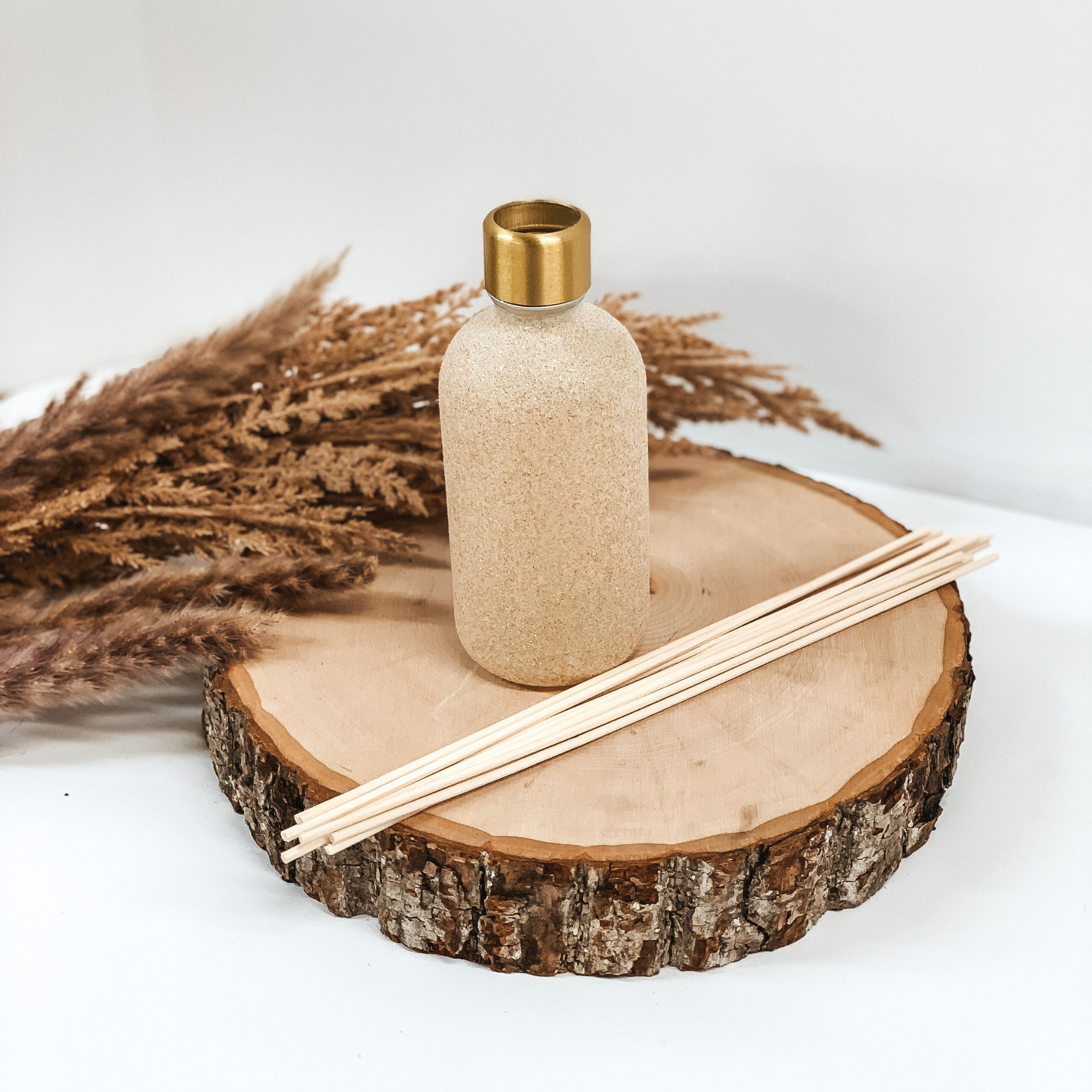 Champagne glitter diffuser bottle pictured on a piece of wood with brown pompous grass in behind the diffuser on a white background. 