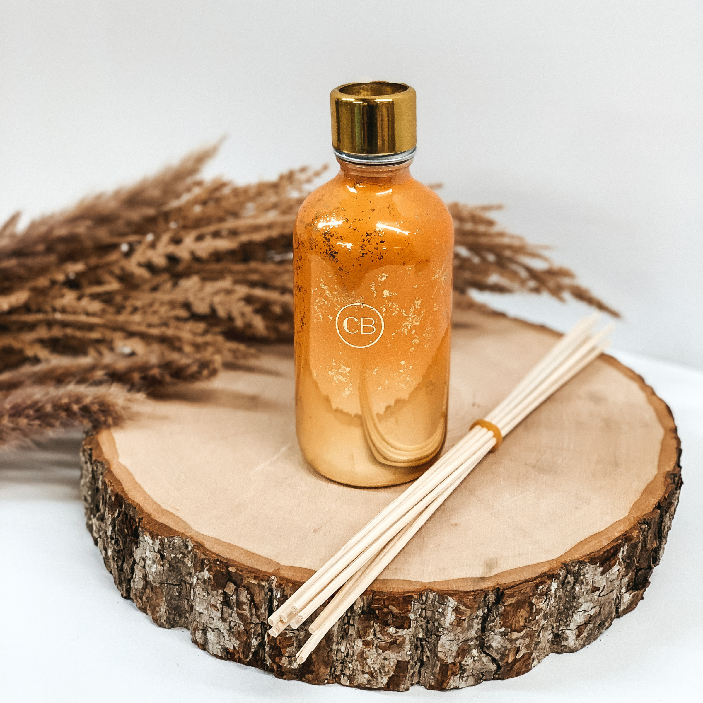 Orange diffuser bottle with gold flecks pictured on a piece of wood with brown pompous grass in behind the diffuser on a white background. 