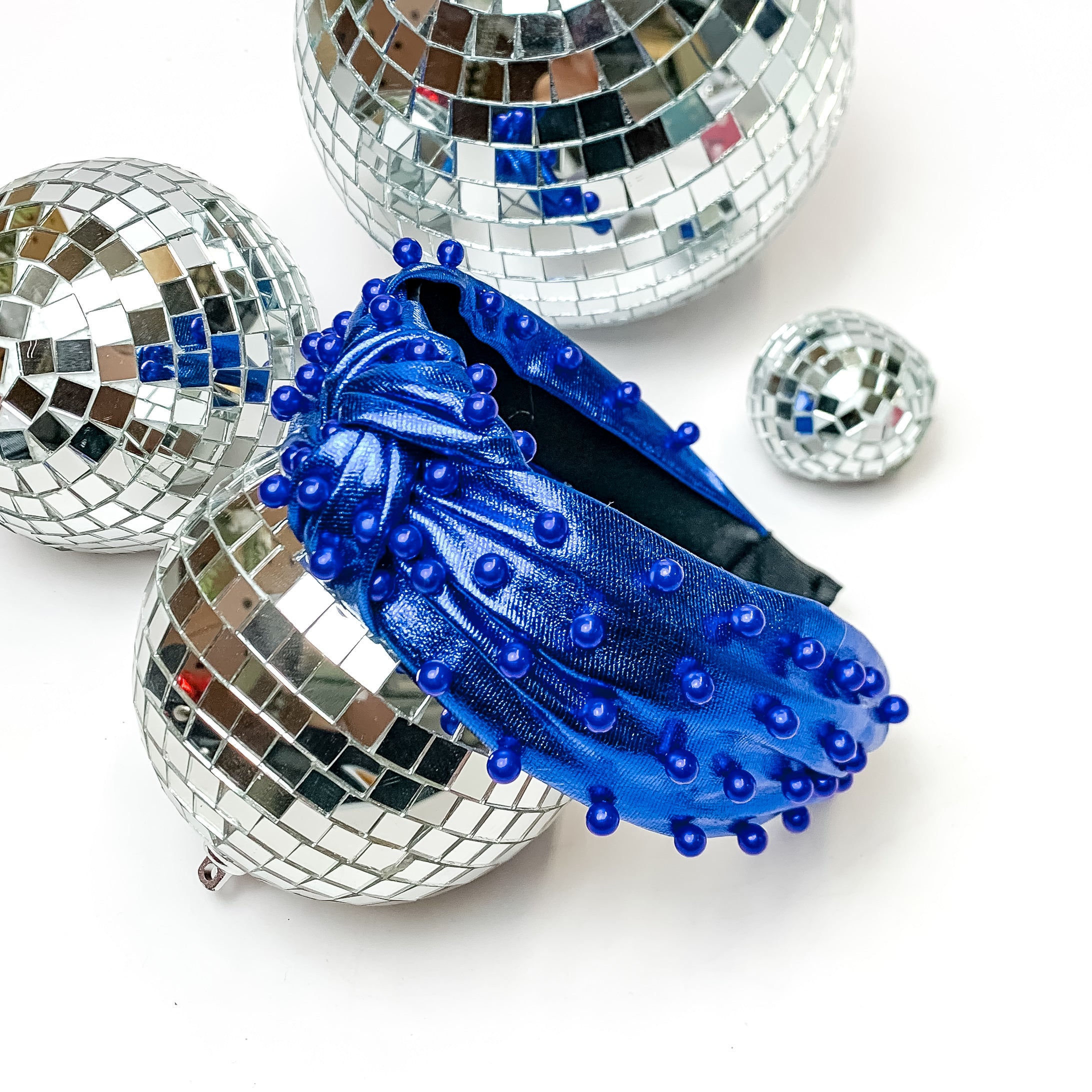 Top of the Charts Blue Headband With Blue Pearls. This headband is pictured on a white background and is surrounded by disco balls.