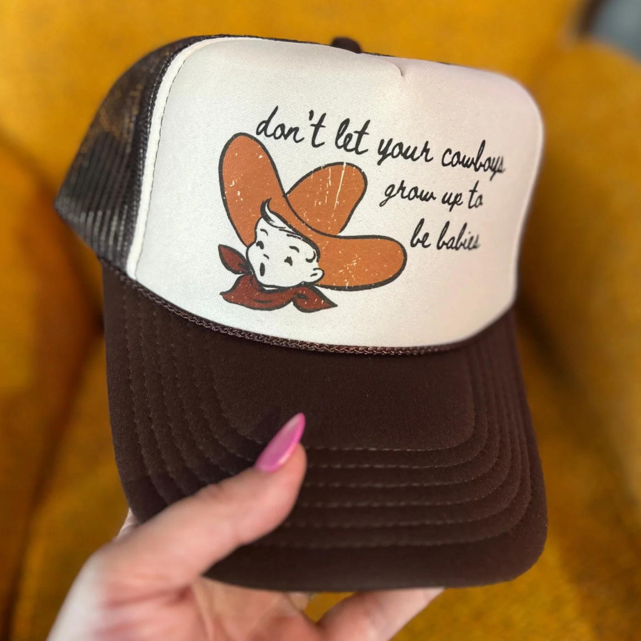 Photo features a brown and cream trucker hat with "don't let your cowboys grow up to be babies" on the front paired with a picture of a baby with a cowboy hat.