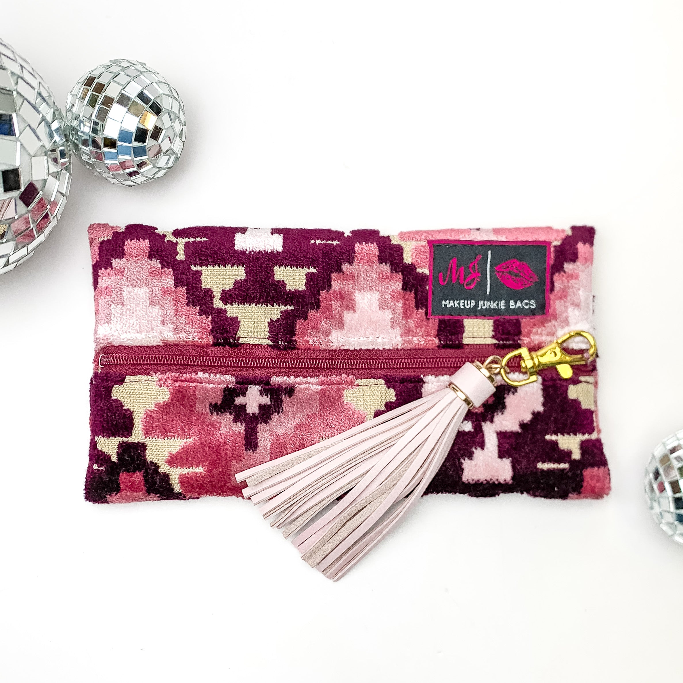 Pictured on a white background with disco balls at top is a mini lay flat bag in a maroon aztec print. This bag includes a middle zipper and a tassel.