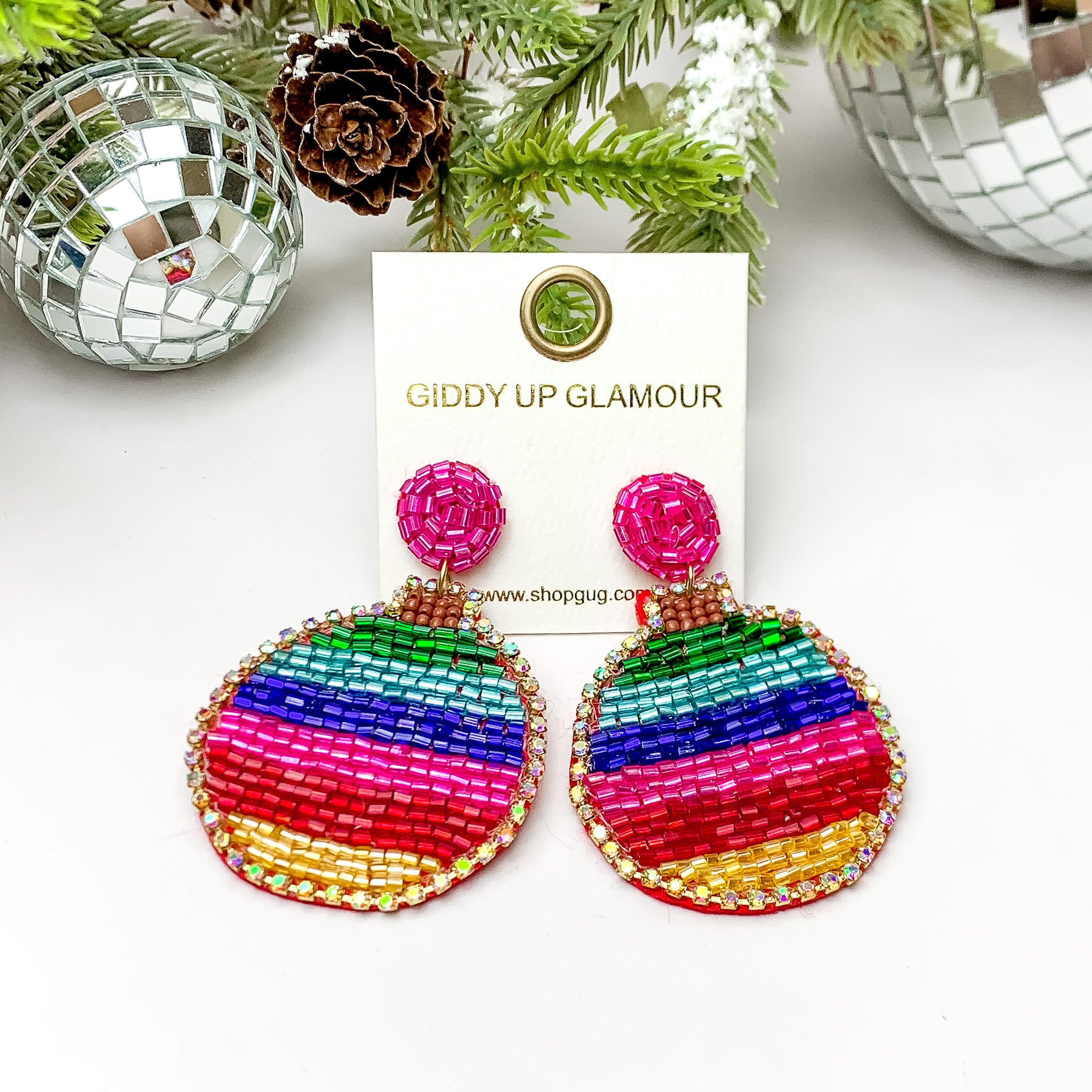 Multicolor striped beaded ornament shaped earrings. These earrings are pictured on a white background with pine trees at the top of the picture. 
