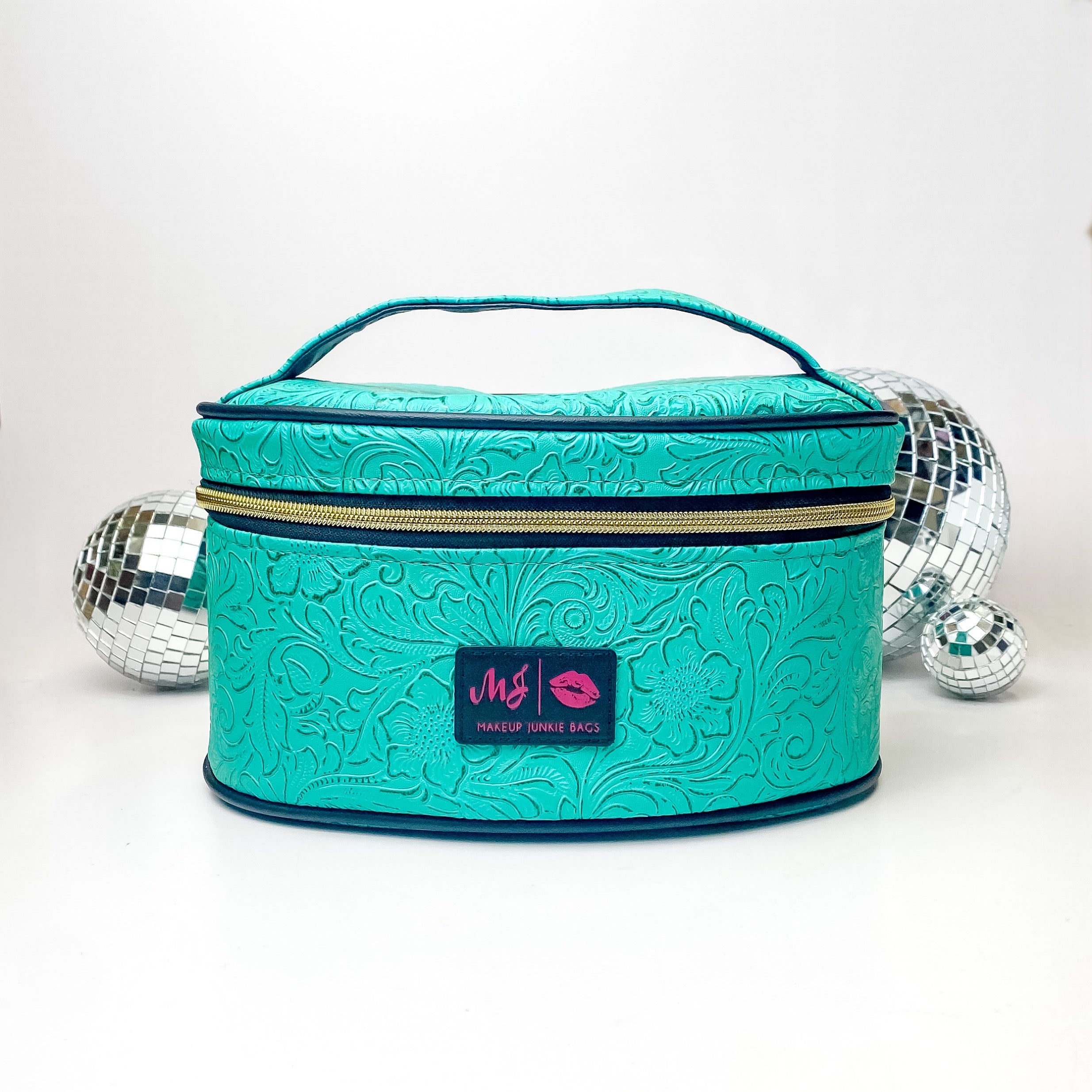 Pictured on a white background with disco balls in the background is a traincase with a top handle in a turquoise leather tooled print. This bag includes a middle zipper and a tassel.