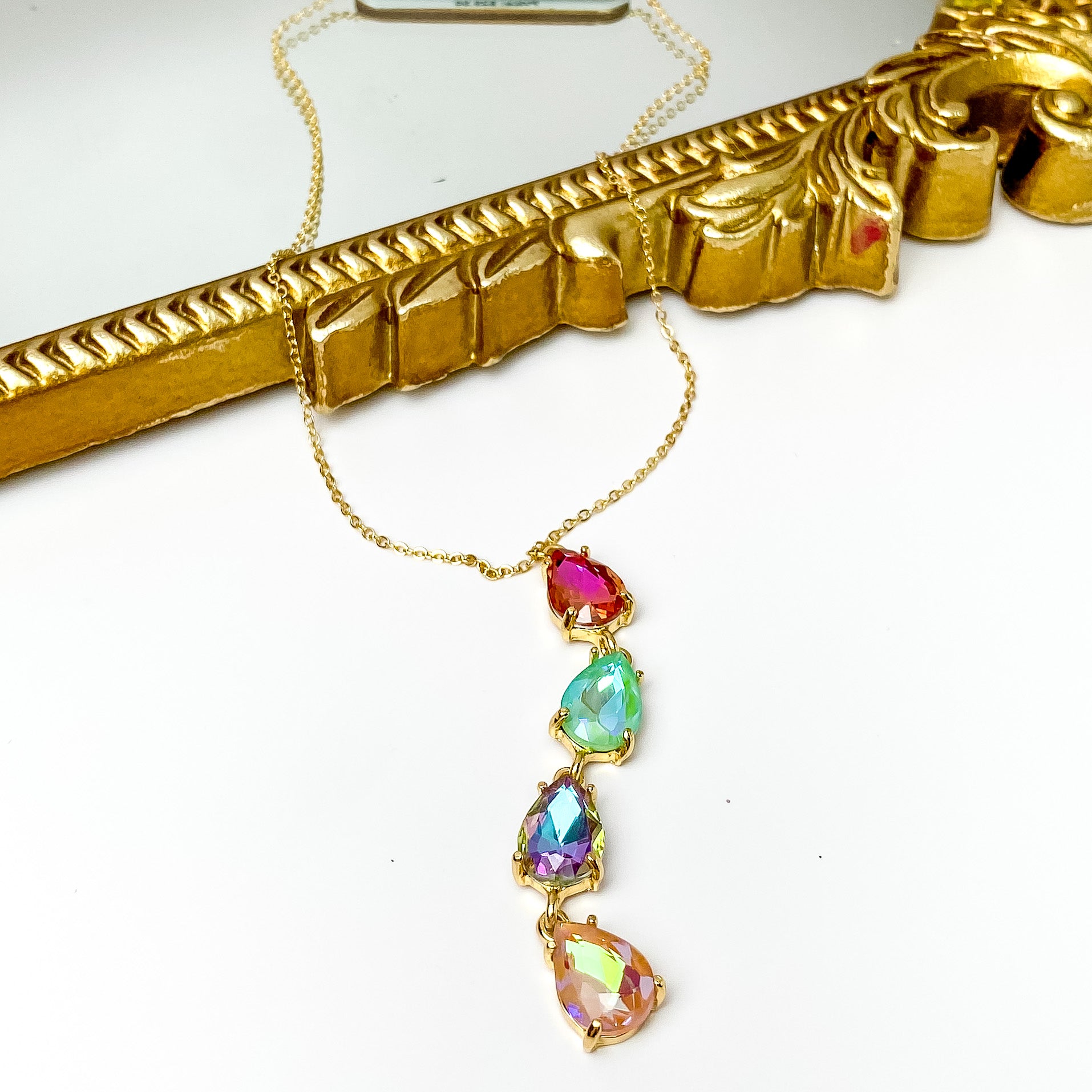 Gold chain necklace with a four crystal drop. This drop are teardrop crystals and include the colors pink, mint, purple, and light pink. This necklace is pictured partially laying on a gold mirror on a white background. 