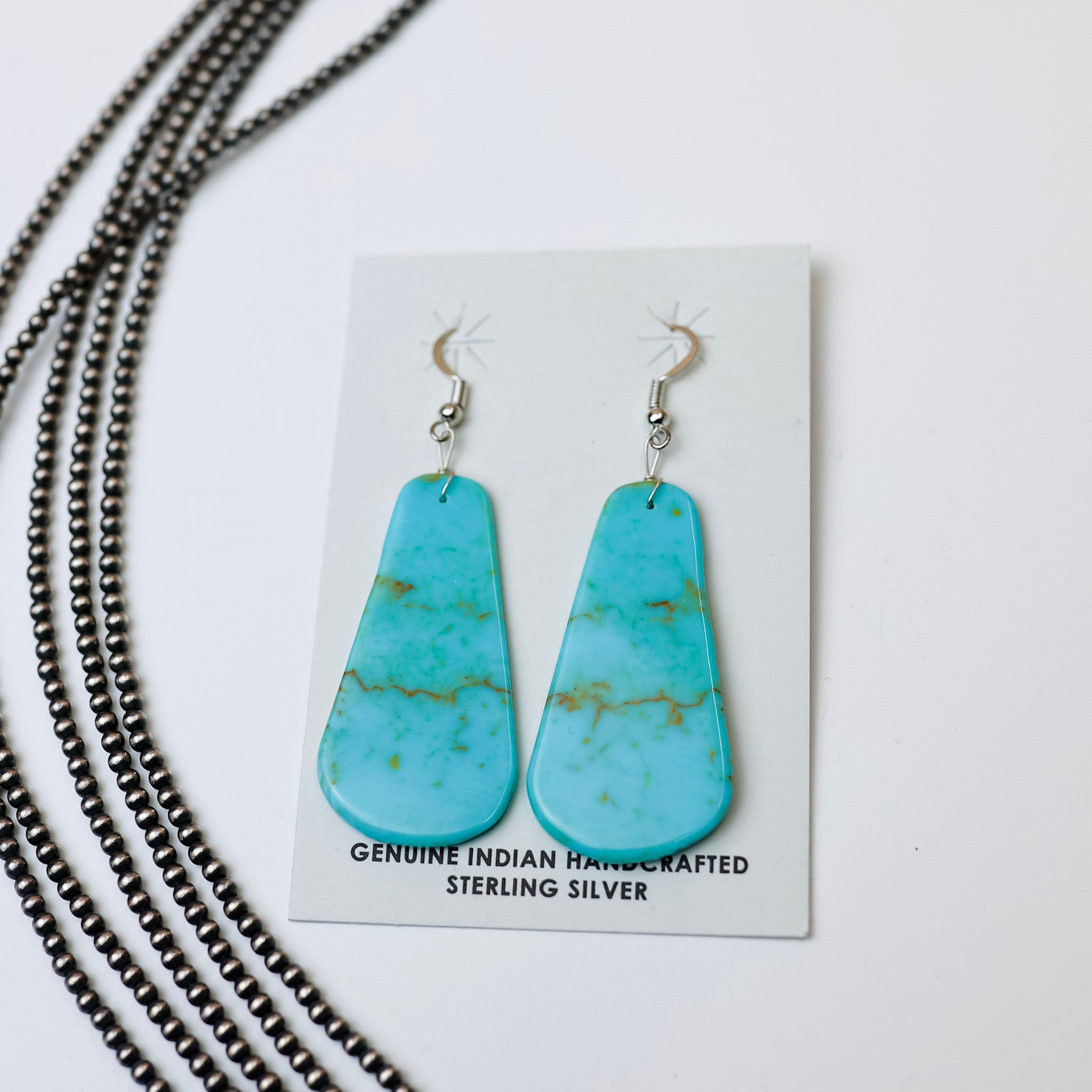 Turquoise slab earrings are placed in the center of the picture. Along the left side of the picture is navajo pearls. All on a white background. 