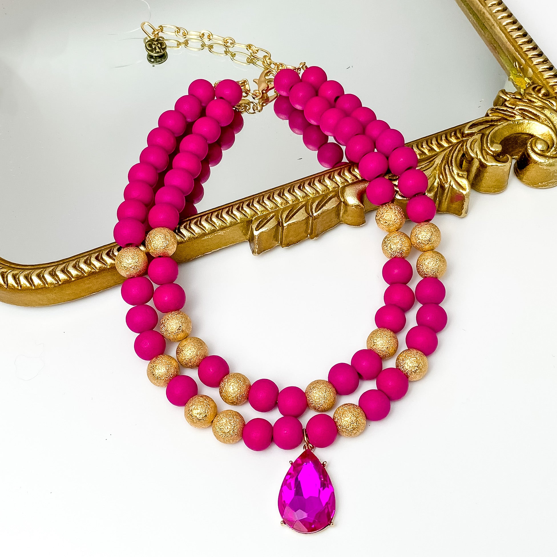 Two strand beaded necklace with a large fuchsia crystal teardrop pendant. This necklace includes fuchsia and gold beads. This necklace is pictured partially laying on a gold mirror on a white background. 