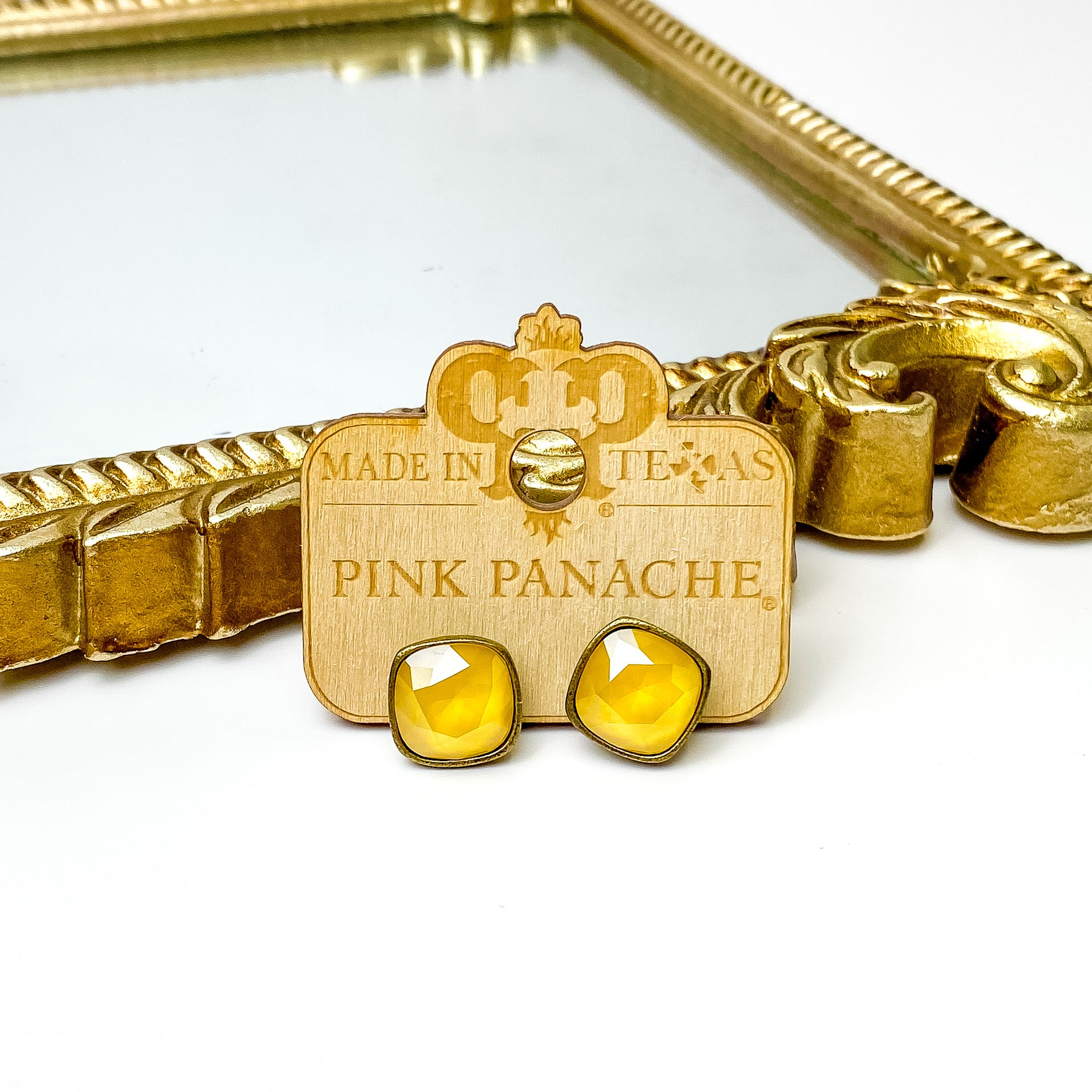 Yellow cushion cut crystal stud earrings with a bronze setting. These earrings are pictured on a Pink Panache wood holder in front of a gold mirror and on a white background.