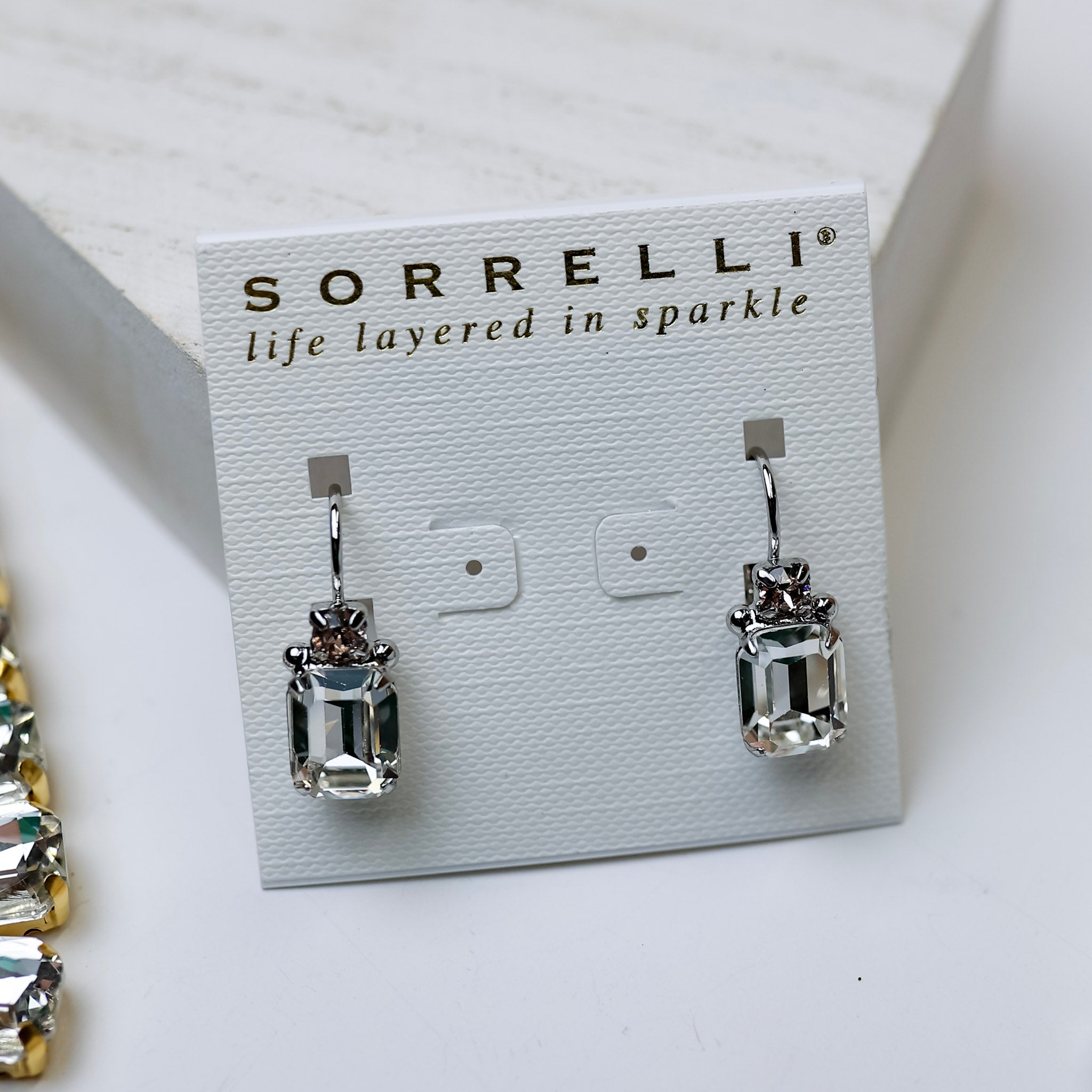 A pair of emerald cut dangle earrings that are clear and silver-tone. Pictured on a white background with crystal necklaces.