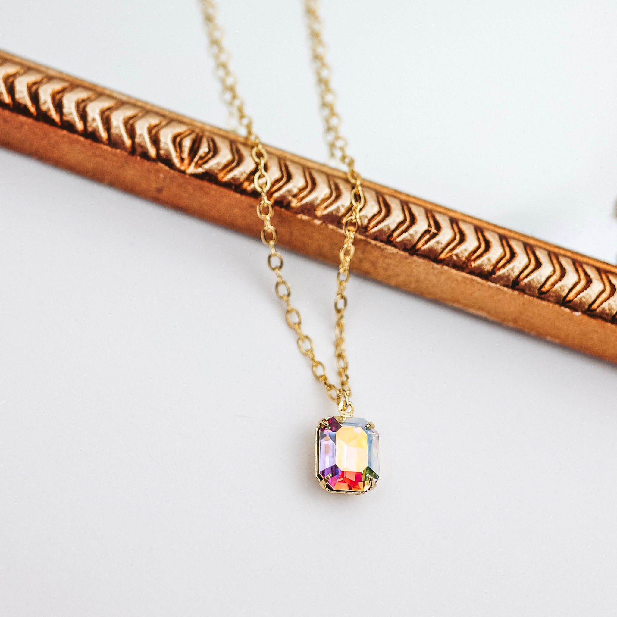 A gold chain necklace with a single colorful crystal pendant that is a square cut. This necklace is pictured on a white background with a mirror.