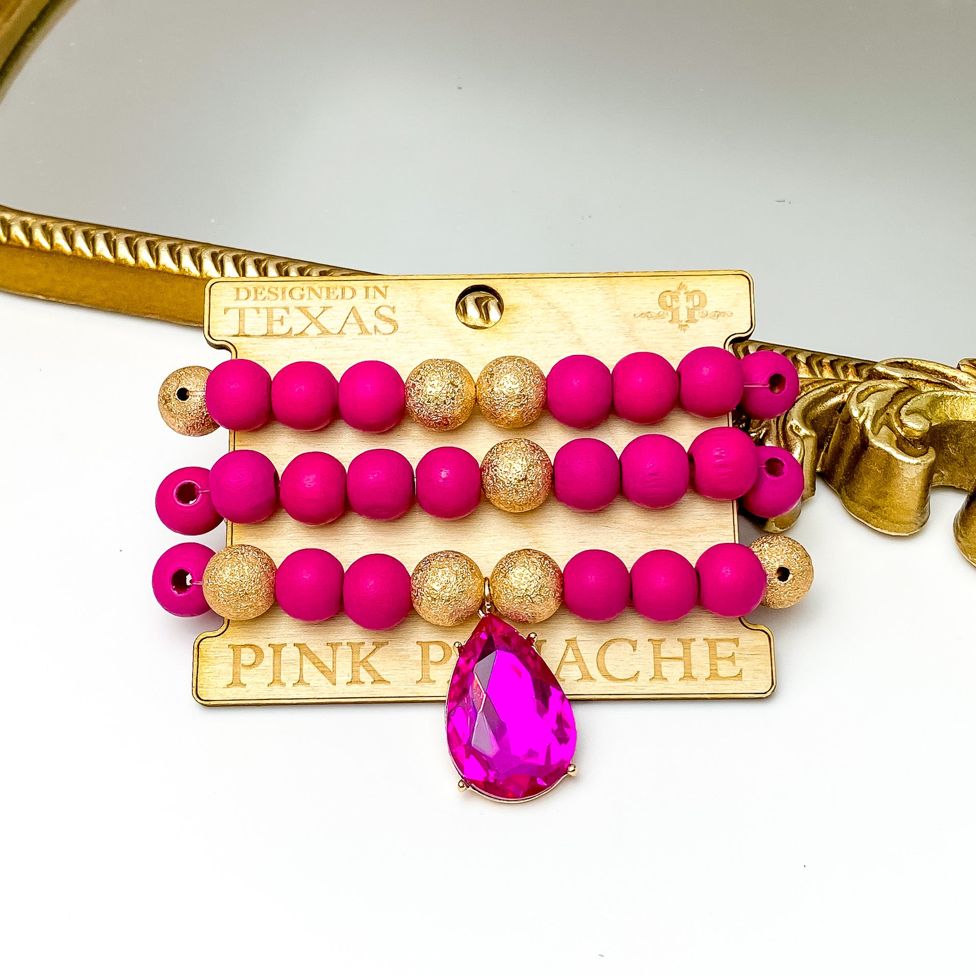 Set of three fuchsia and gold bead bracelets. This set also includes a large, fuchsia teardrop crystal. These bracelets are pictured on a Pink Panache wood holder in front of a gold mirror and on a white background.