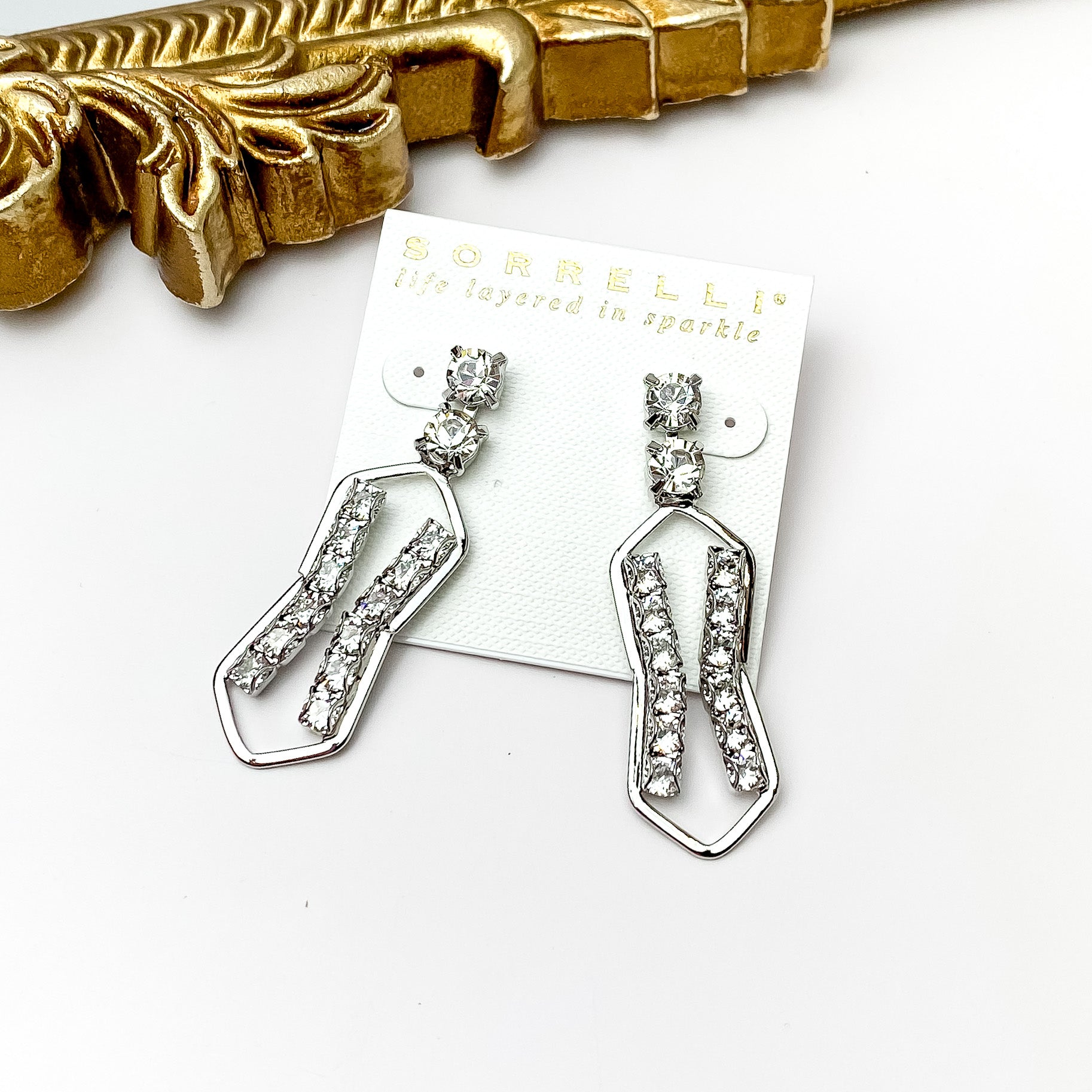 Two clear crystals on a post back with a silver, hourglass shaped drop. The gold drop include a line of clear crystals on each side. These earrings are pictured in front of a gold mirror on a white background. 