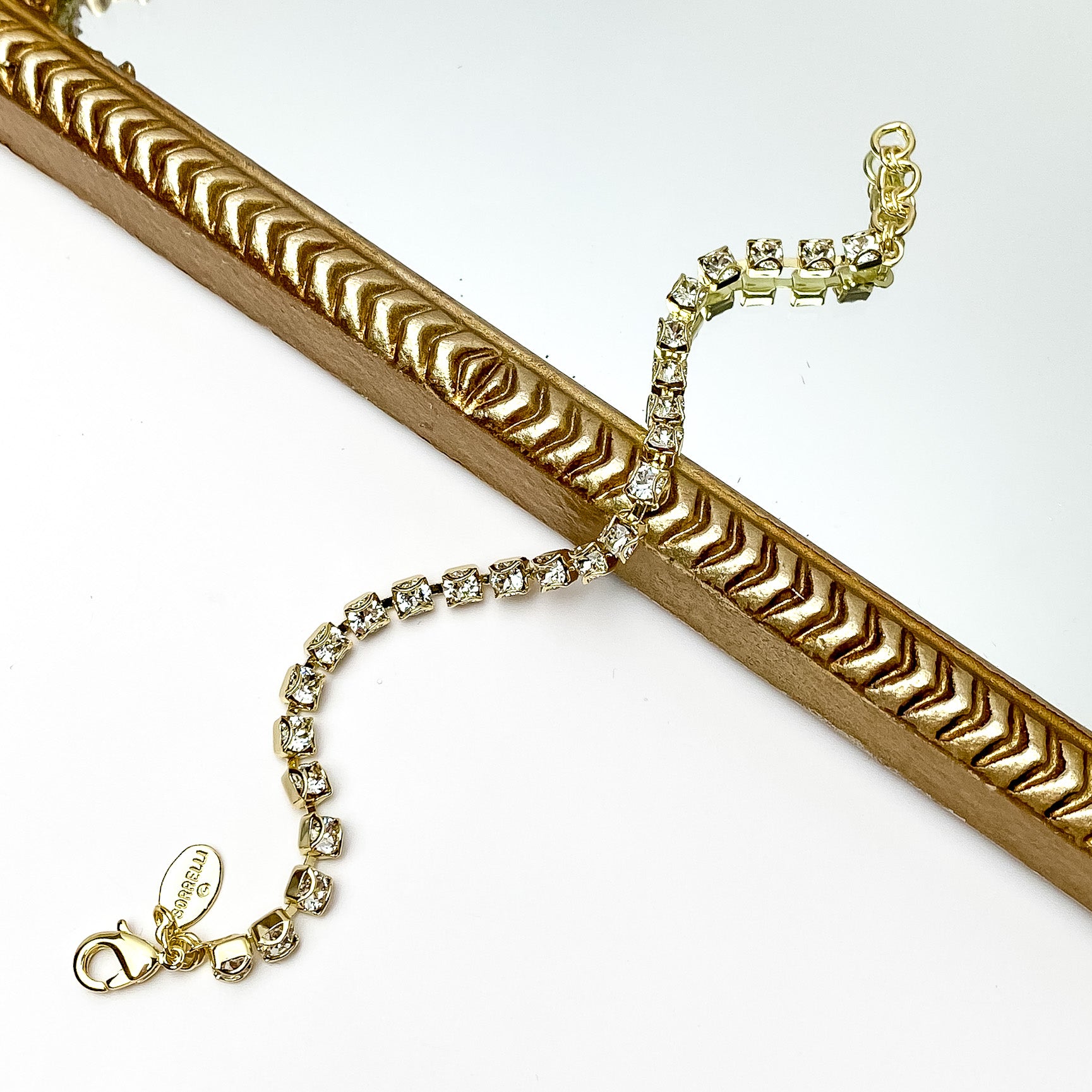 Gold tennis bracelet with clear crystals. Pictured on a white background and a gold frame through the middle.