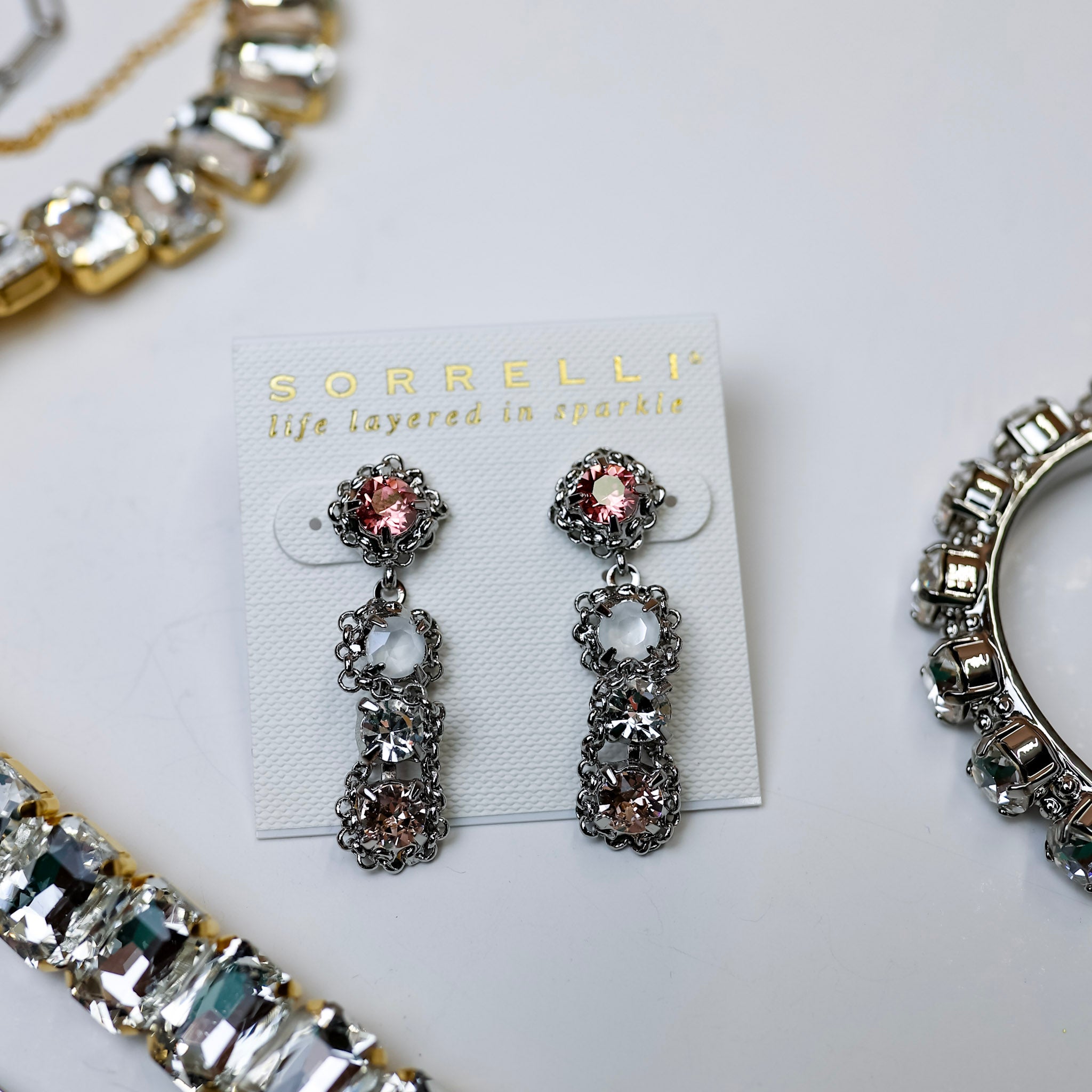 A pair of silver-tone dangle earrings with blush pink and white crystals and chain detailing. Pictured on a white background with crystal necklaces.