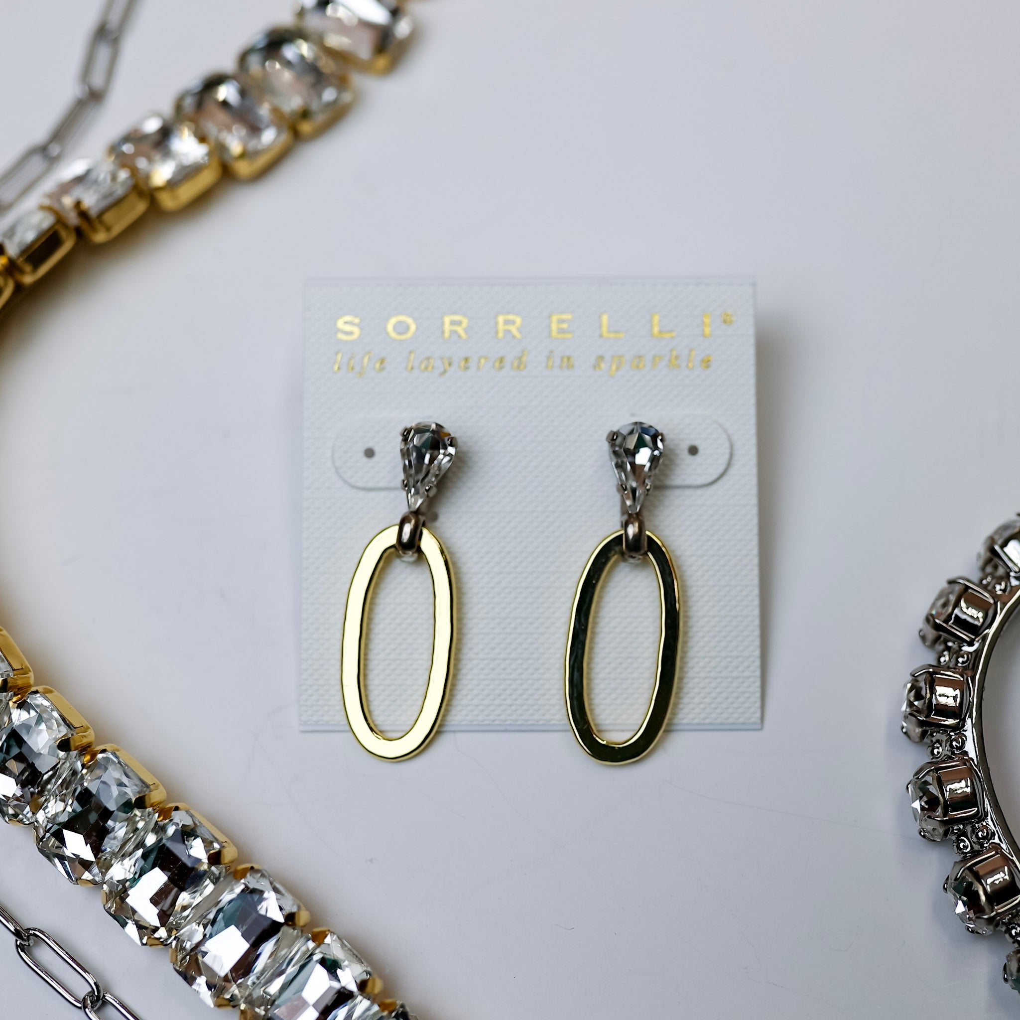 A pair of crystal post earrings with a gold tone oval dangle. Pictured on a white background with crystal necklaces.