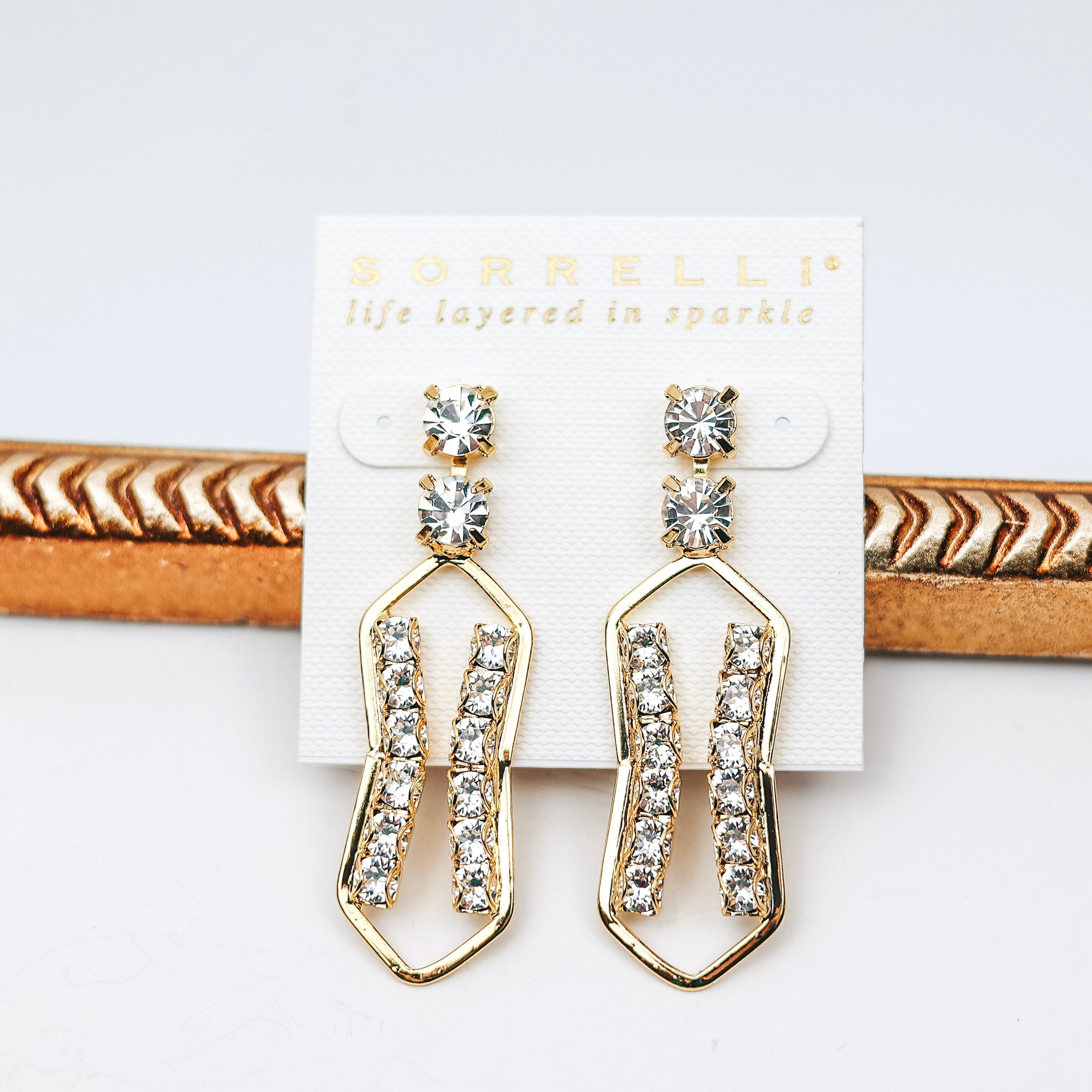A pair of gold dangle earrings with clear crystals all over. These earrings are pictured on a white background.