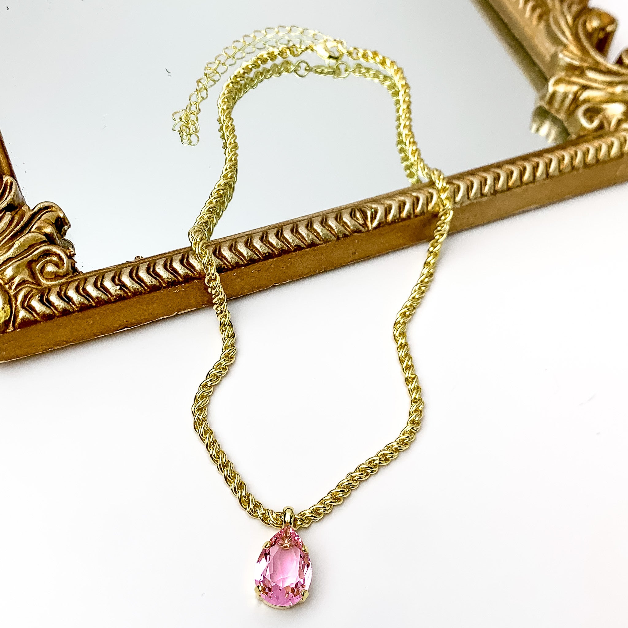 Pictured is a gold chain necklace with a pink teardrop crystal charm. This necklace is pictured partially laying on a gold mirror on a white background.