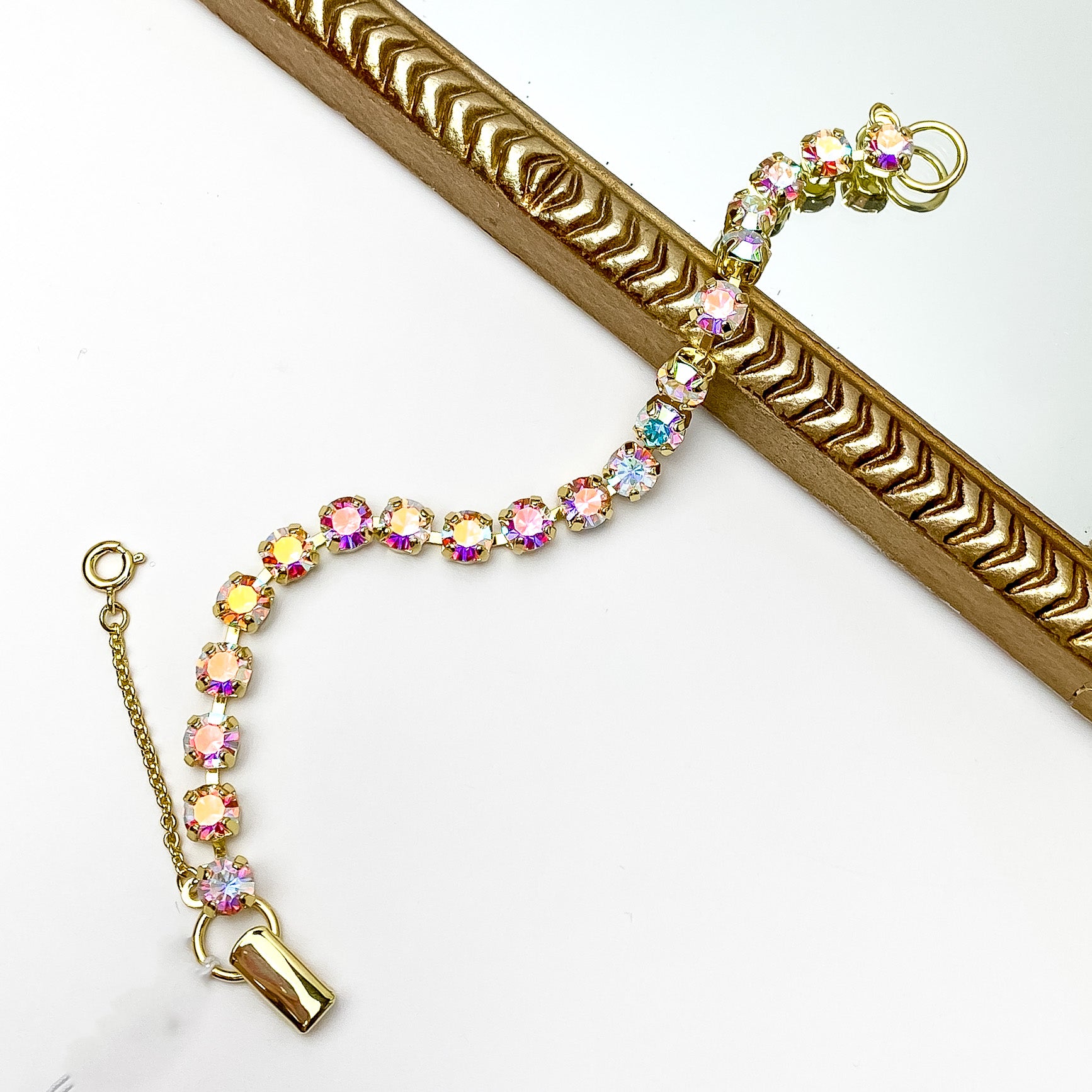 Gold tennis bracelet with Aurora Borealis crystals. Pictured on a white background with a gold frame through the middle. 