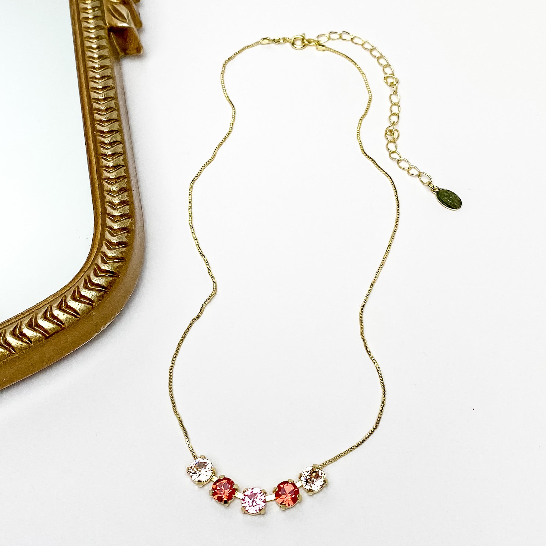 Pictured is a gold necklace with a five crystals. The center crystal is pink, the next one on each side is red, and finally the last two are a light champagne colored crystal. This necklace is pictured on a white background with a gold mirror on the left side.