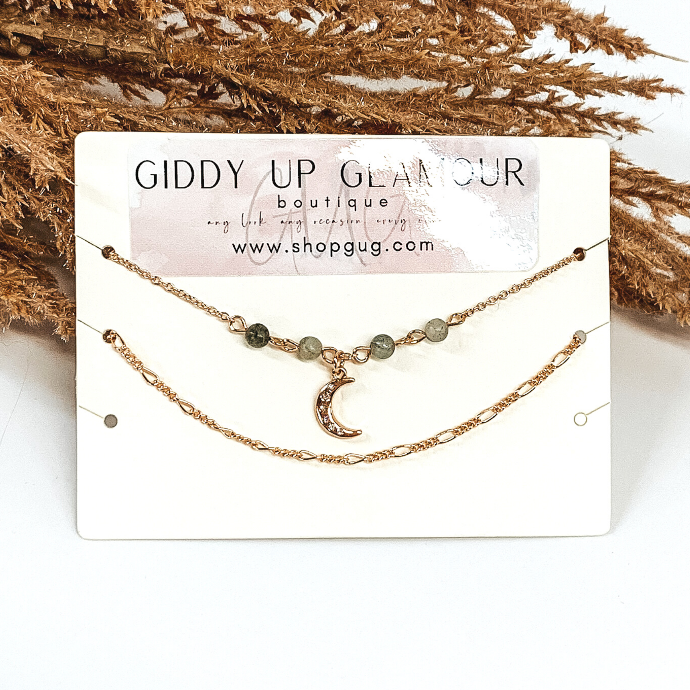 Two gold chain anklets. One was a plain anklet, while the second anklet has 4 grey beads with a gold moon charm in the center that has crystals on it. They are pictured on a white background that has brown floral. 