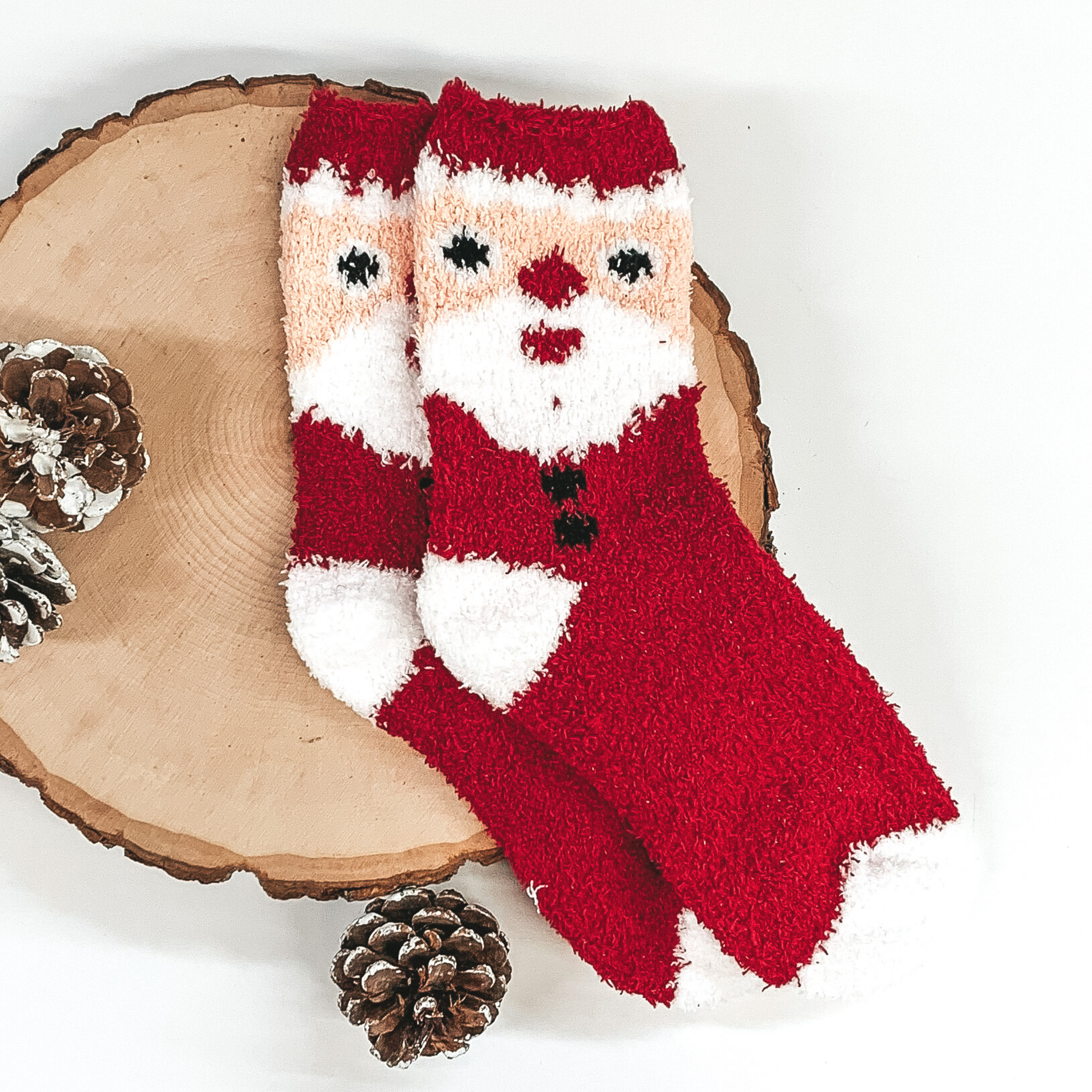 Fuzzy red socks with a colored santa face on the ankle part of the socks. These socks are pictured laying on a piece of wood that is on a white background.