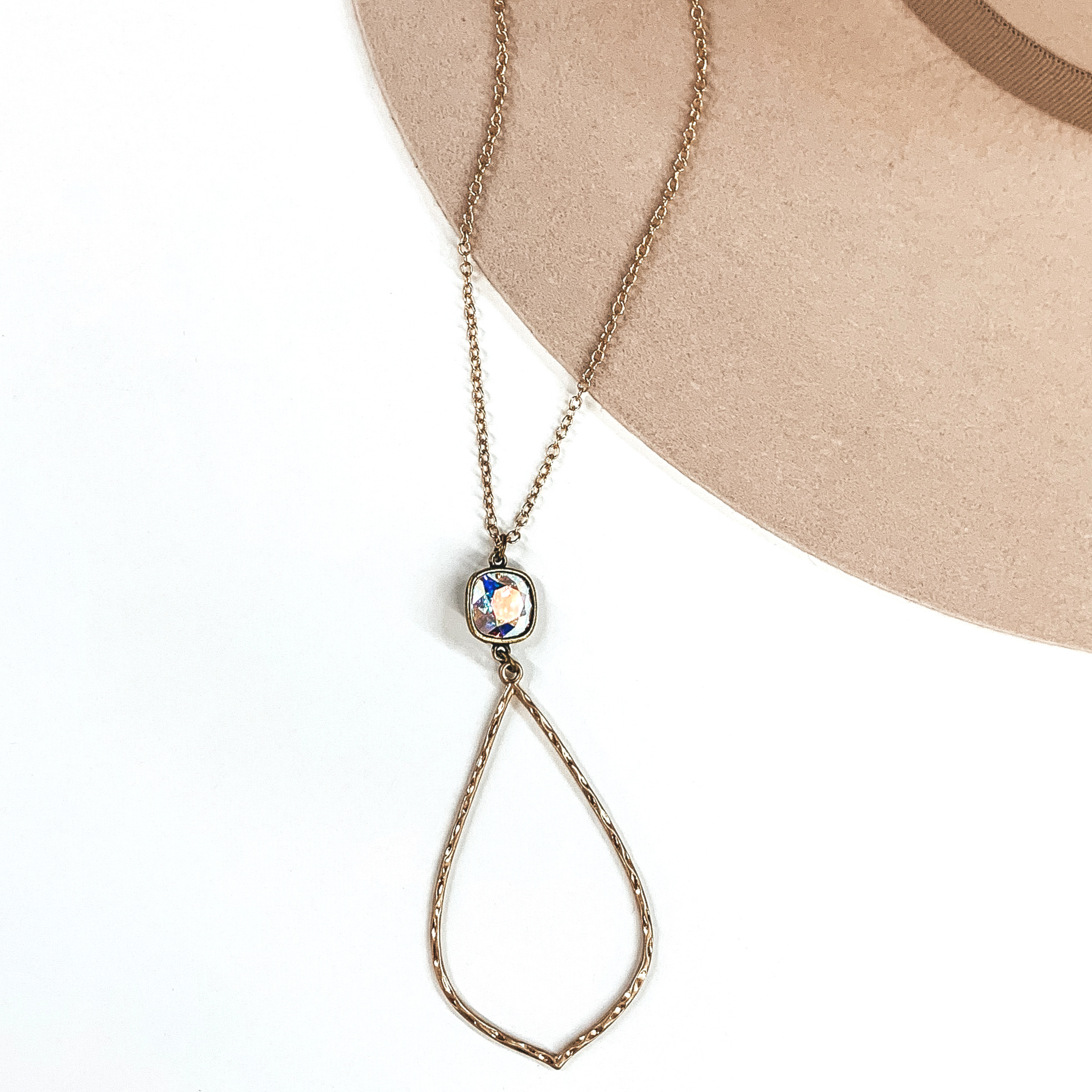 Small gold chained necklace with hanging ab cushion cut crystal and thin hammered teardrop pendant. This necklace is pictured on a white and beige background.