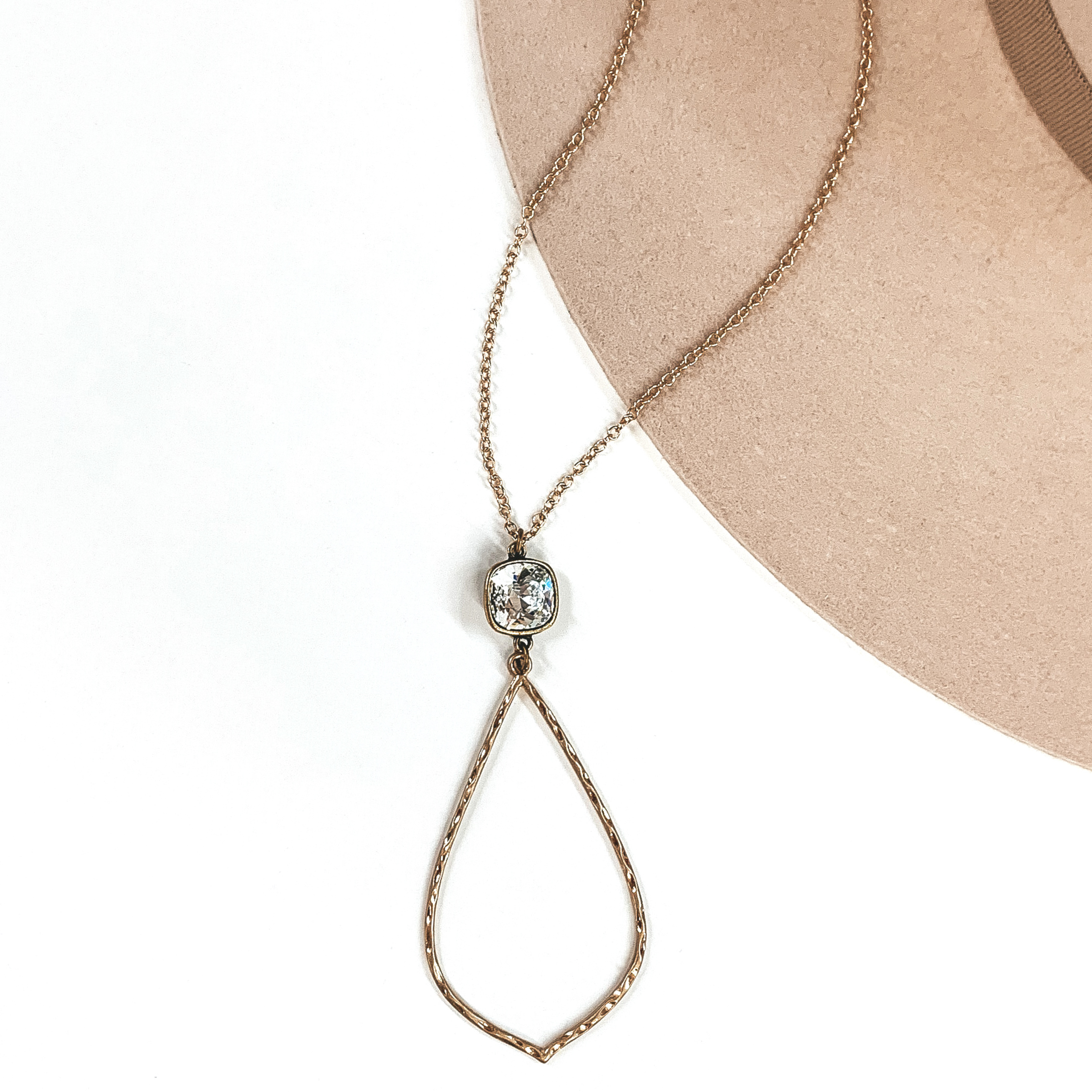 Small gold chained necklace with hanging clear cushion cut crystal and thin hammered teardrop pendant. This necklace is pictured on a white and beige background.