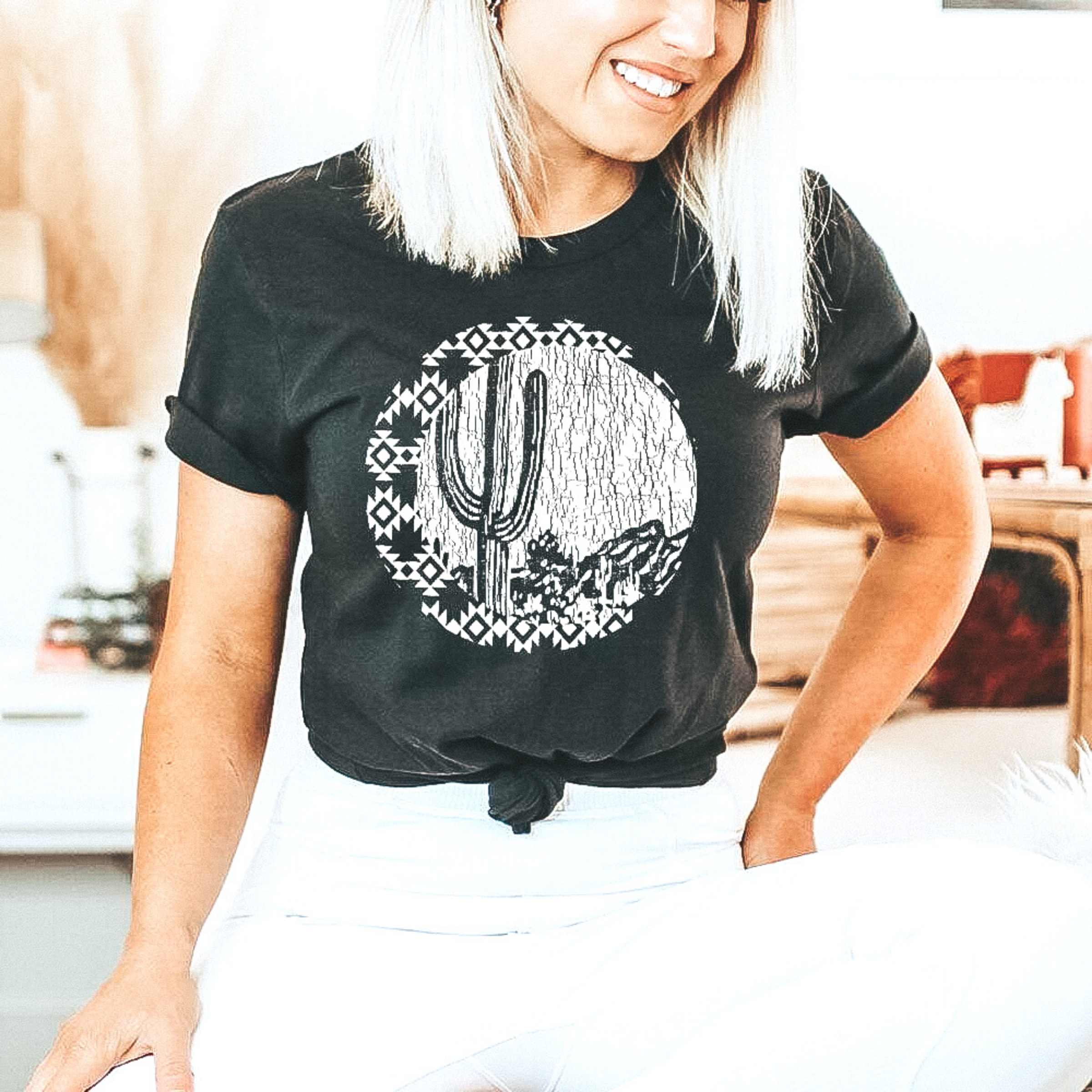 Black graphic tee with saguaro cactus, mountains in background and chipped print skyline.  The graphic has an aztec design surounding. Blonde model has paired back with white leggings.