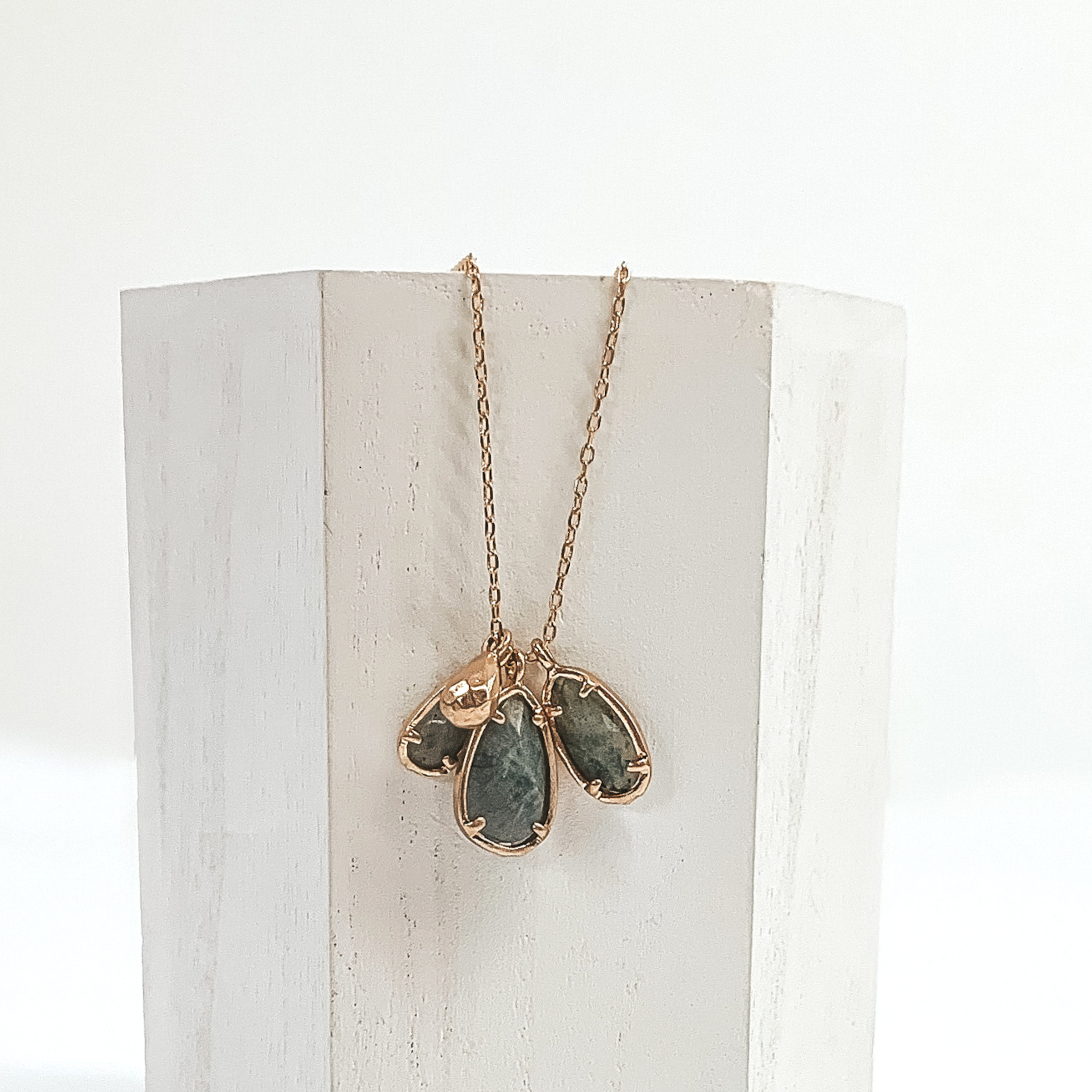 Small gold chain with three grey marbled teardrop stone pendants outlined in gold. It also has a tiny gold teardrop pendant. This necklace is pictured laying a white piece of wood on a white background.