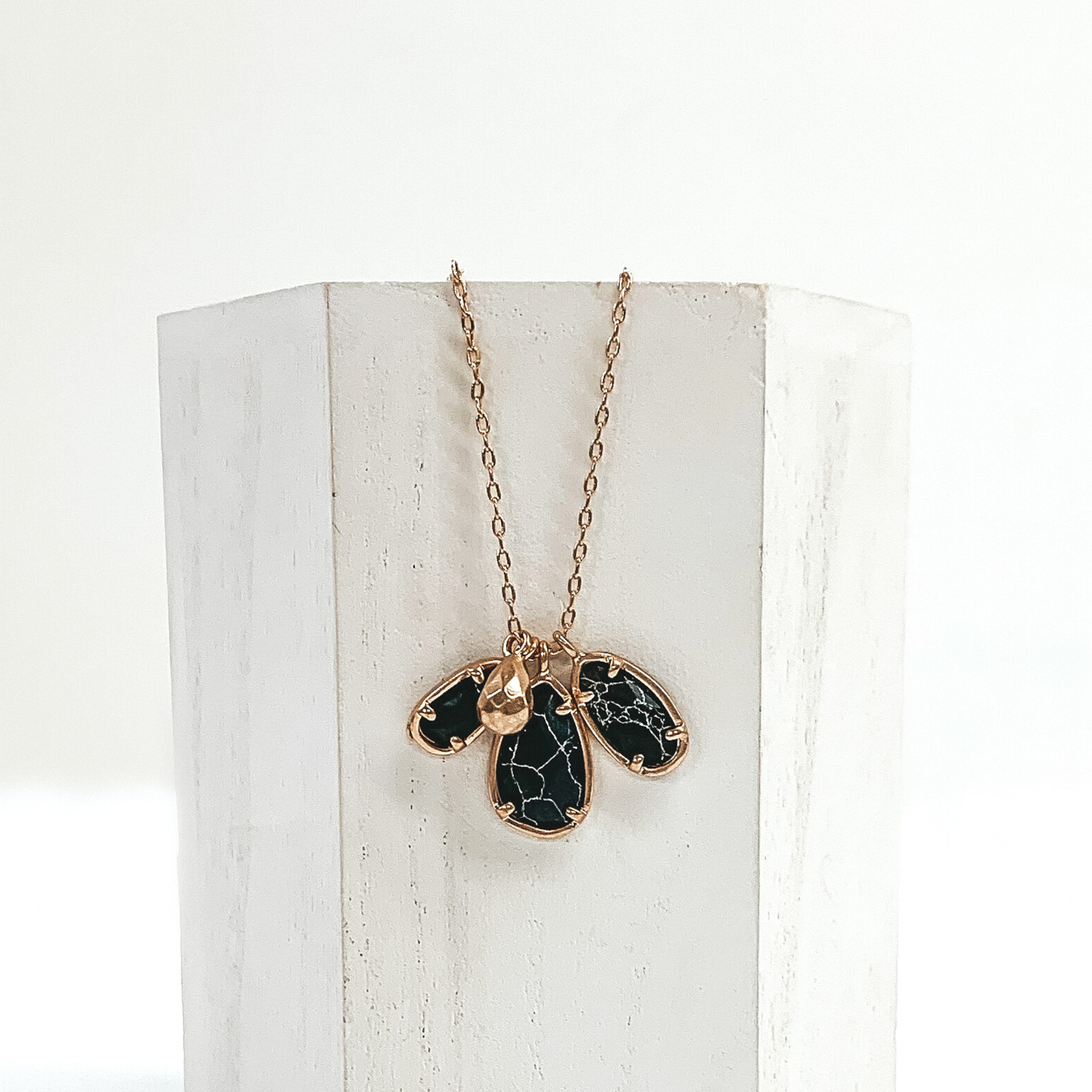 Small gold chain with three black marbled teardrop stone pendants outlined in gold. It also has a tiny gold teardrop pendant. This necklace is pictured laying a white piece of wood on a white background.